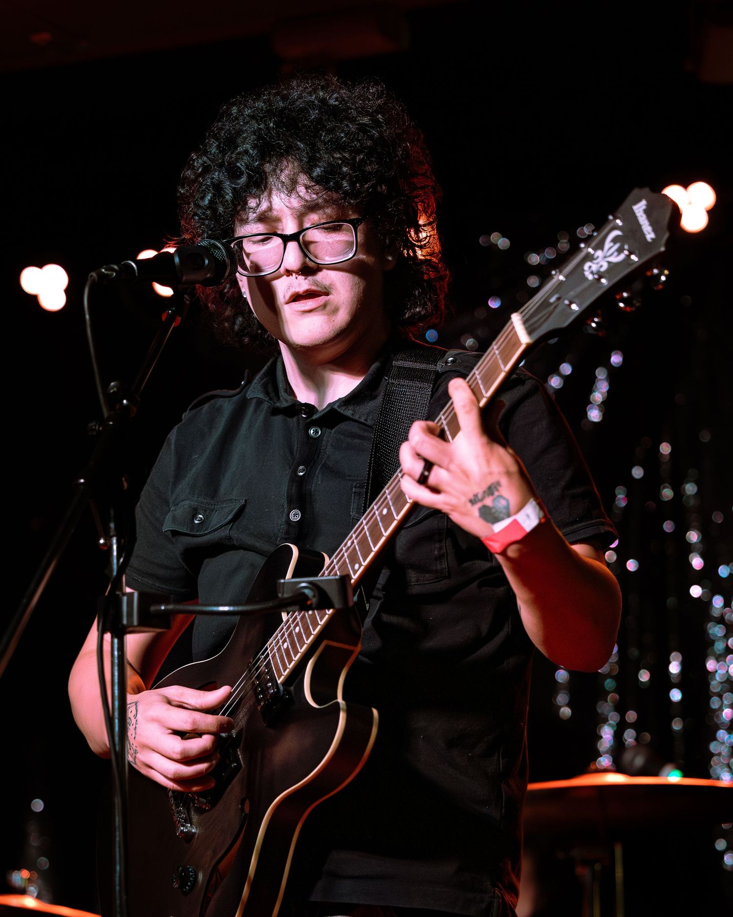 Curly-haired musician with glasses playing acoustic guitar at microphone during live performance.