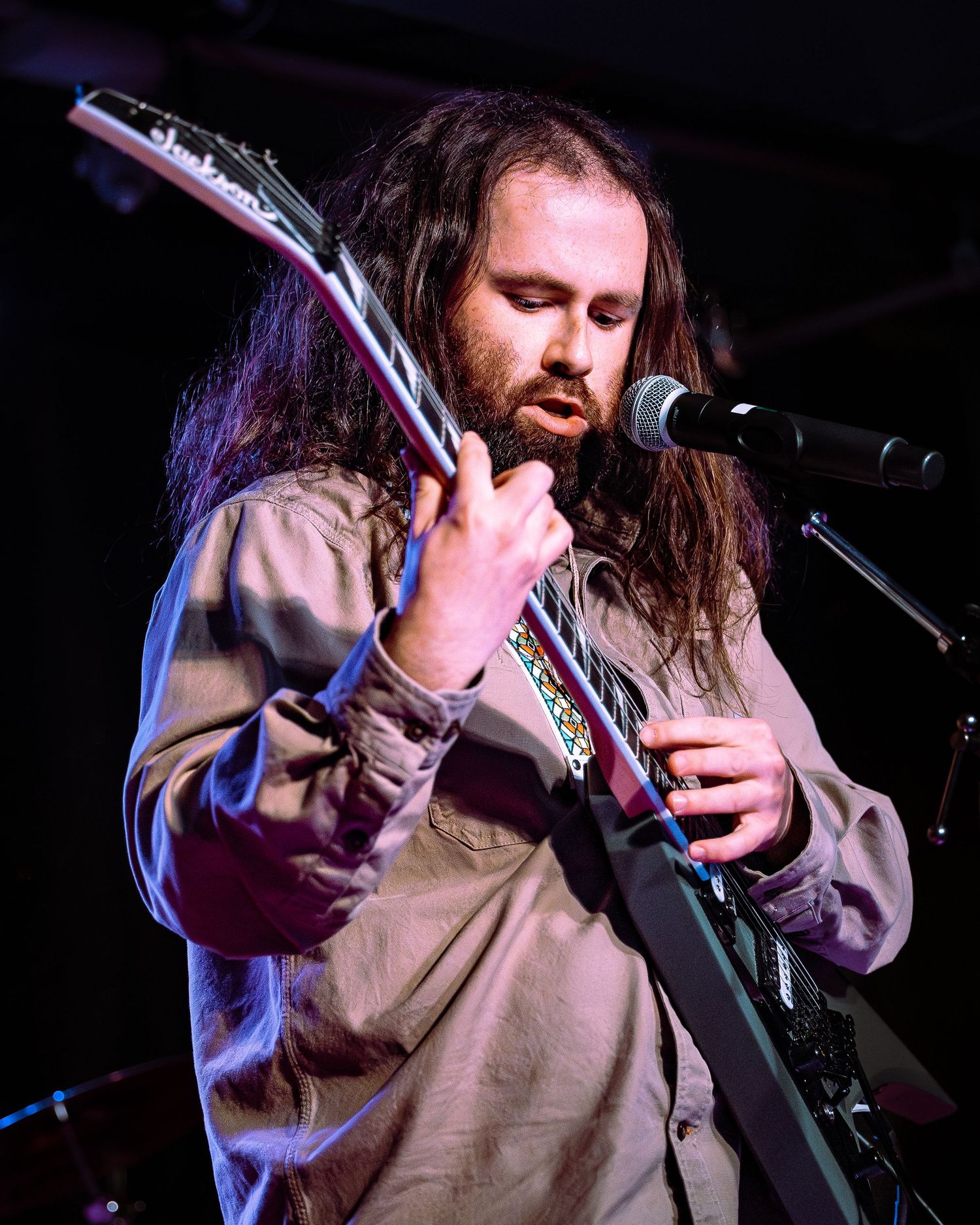 Long-haired bearded guitarist plays dark electric guitar at microphone on stage.