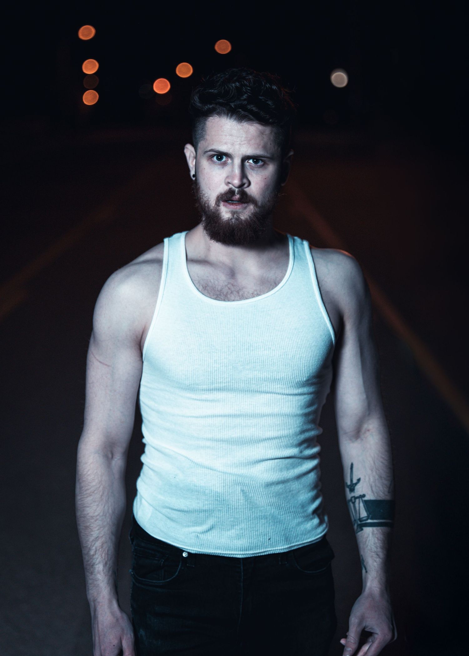 Person in white tank top and dark pants poses against black background.