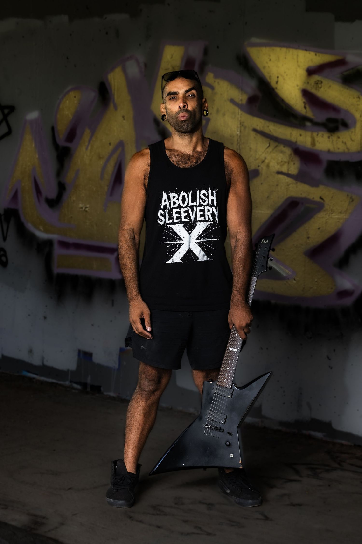 Person in black sleeveless shirt with tattoos poses against graffiti wall in dark urban setting.