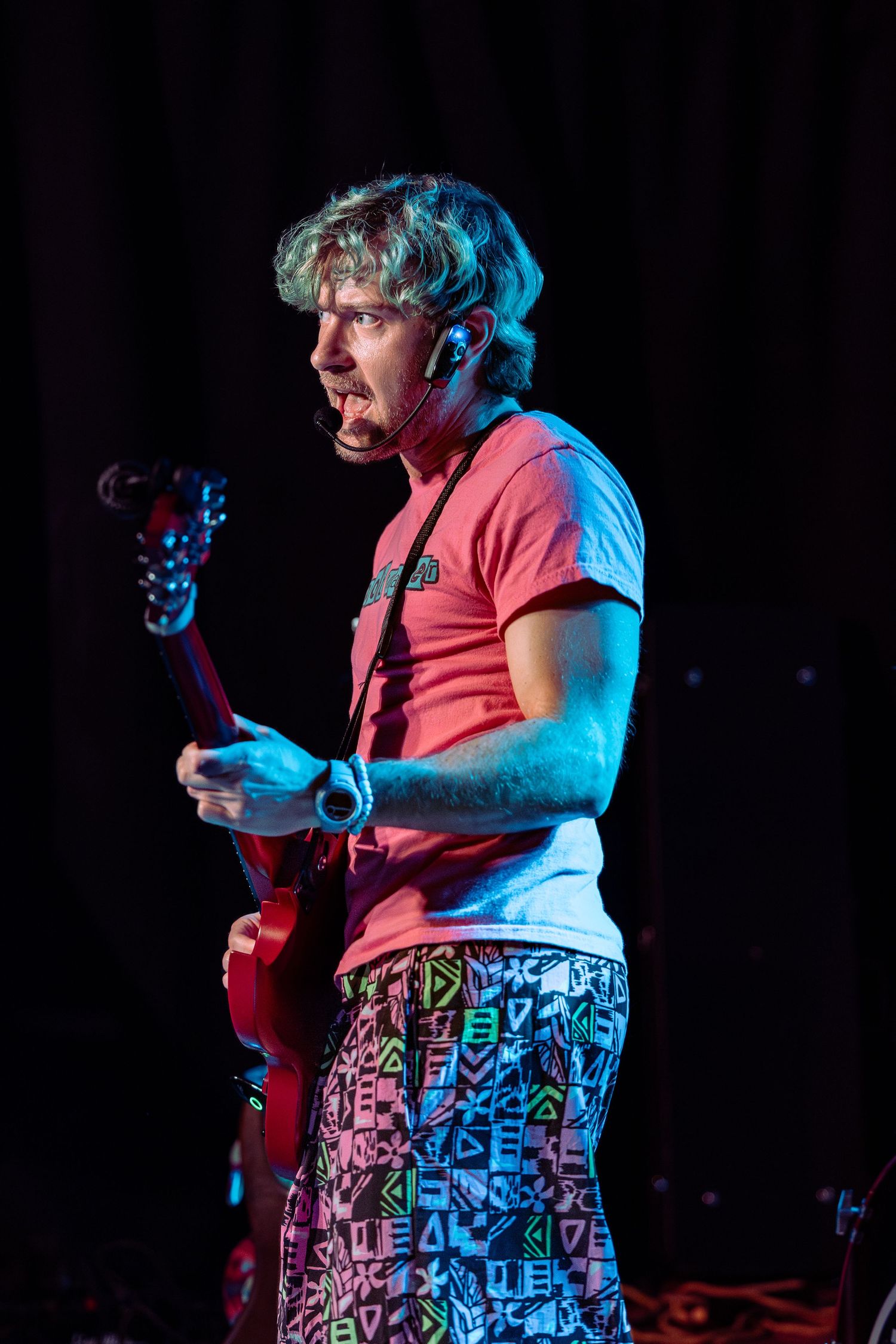 Musician in red t-shirt and colorful shorts plays electric guitar on stage under blue and red lights.
