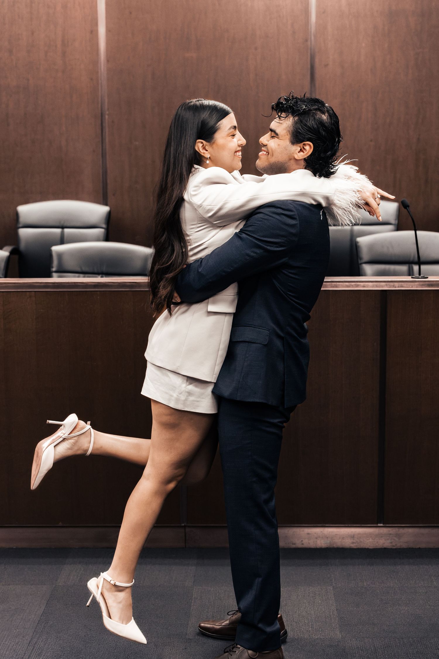 Couple embracing and smiling in a courtroom, woman in white suit lifting leg, man in dark suit, wooden paneling background.