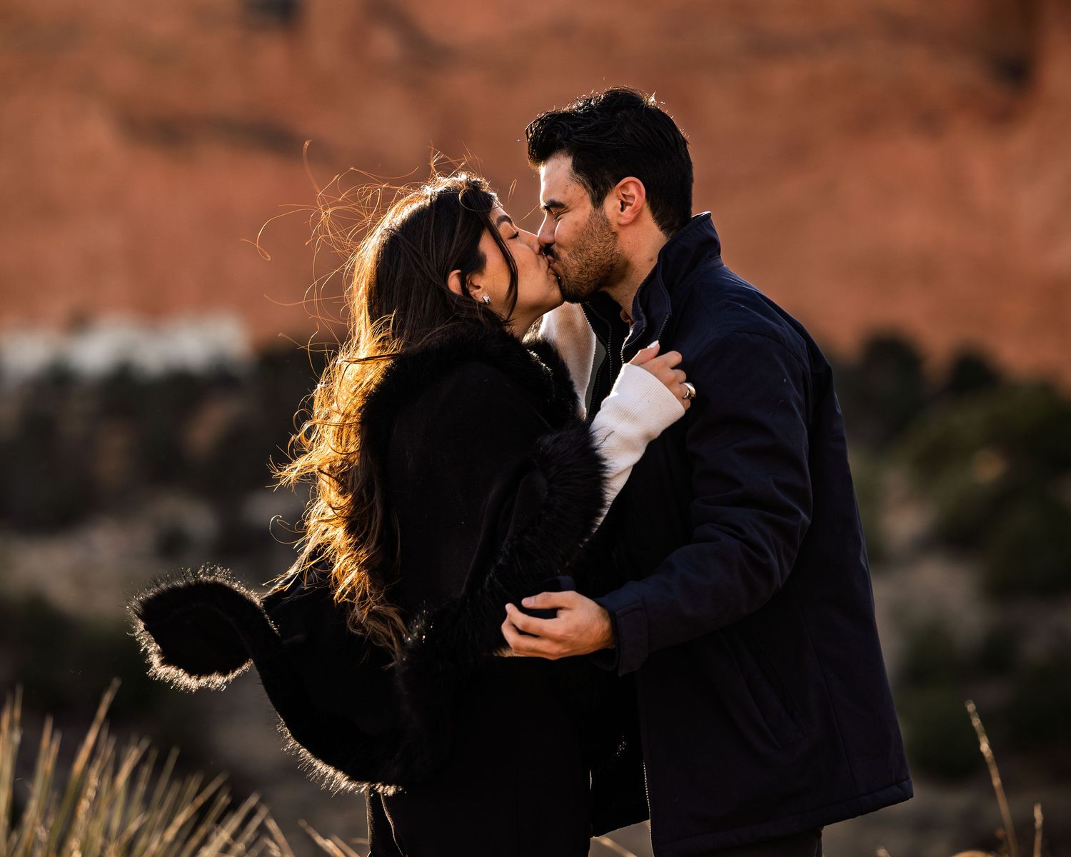 A couple embraces in a romantic kiss at sunset against a backdrop of red desert cliffs.