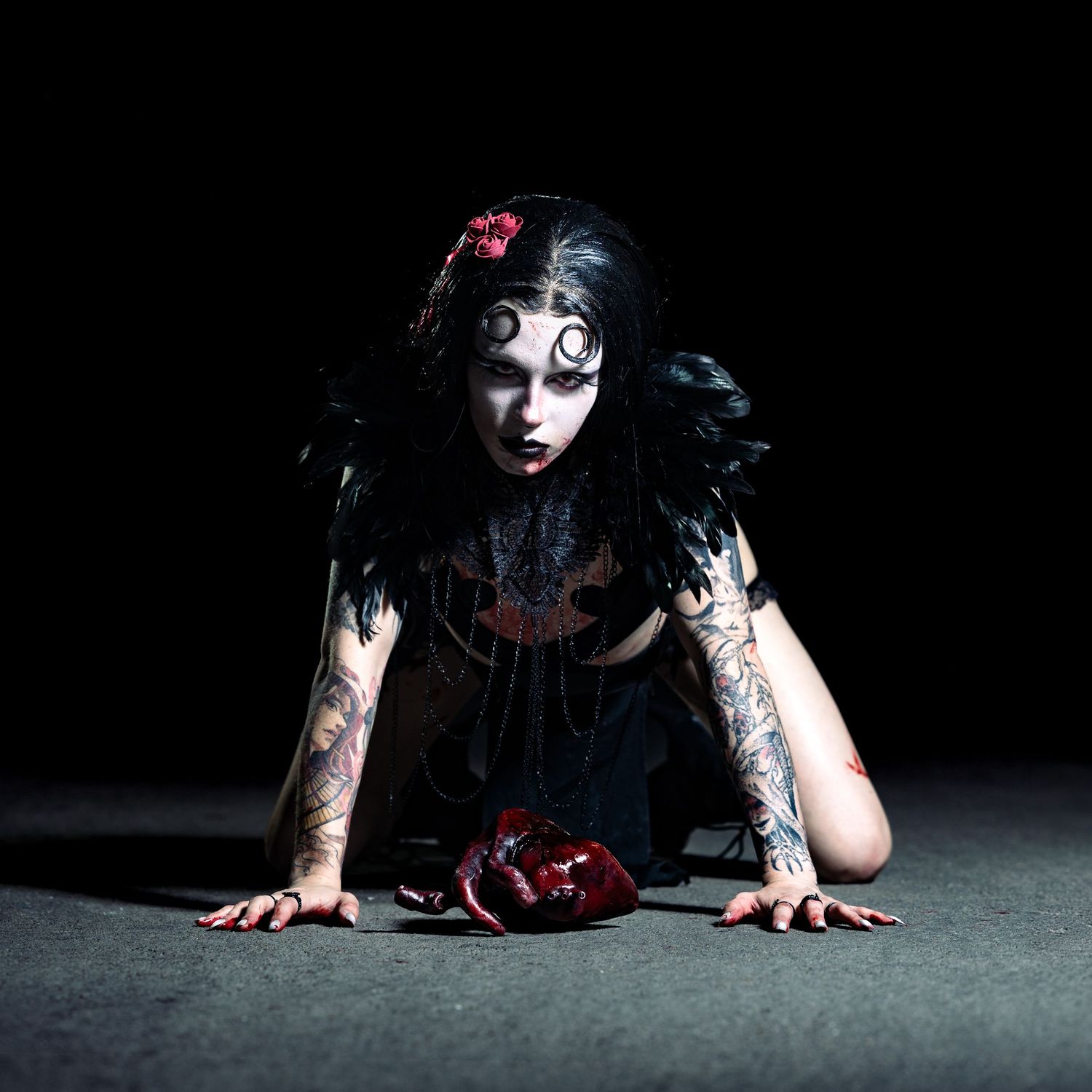 Gothic woman with spiral eyes, black feathers, and tattoos crawls on dark floor in dramatic horror-themed makeup.