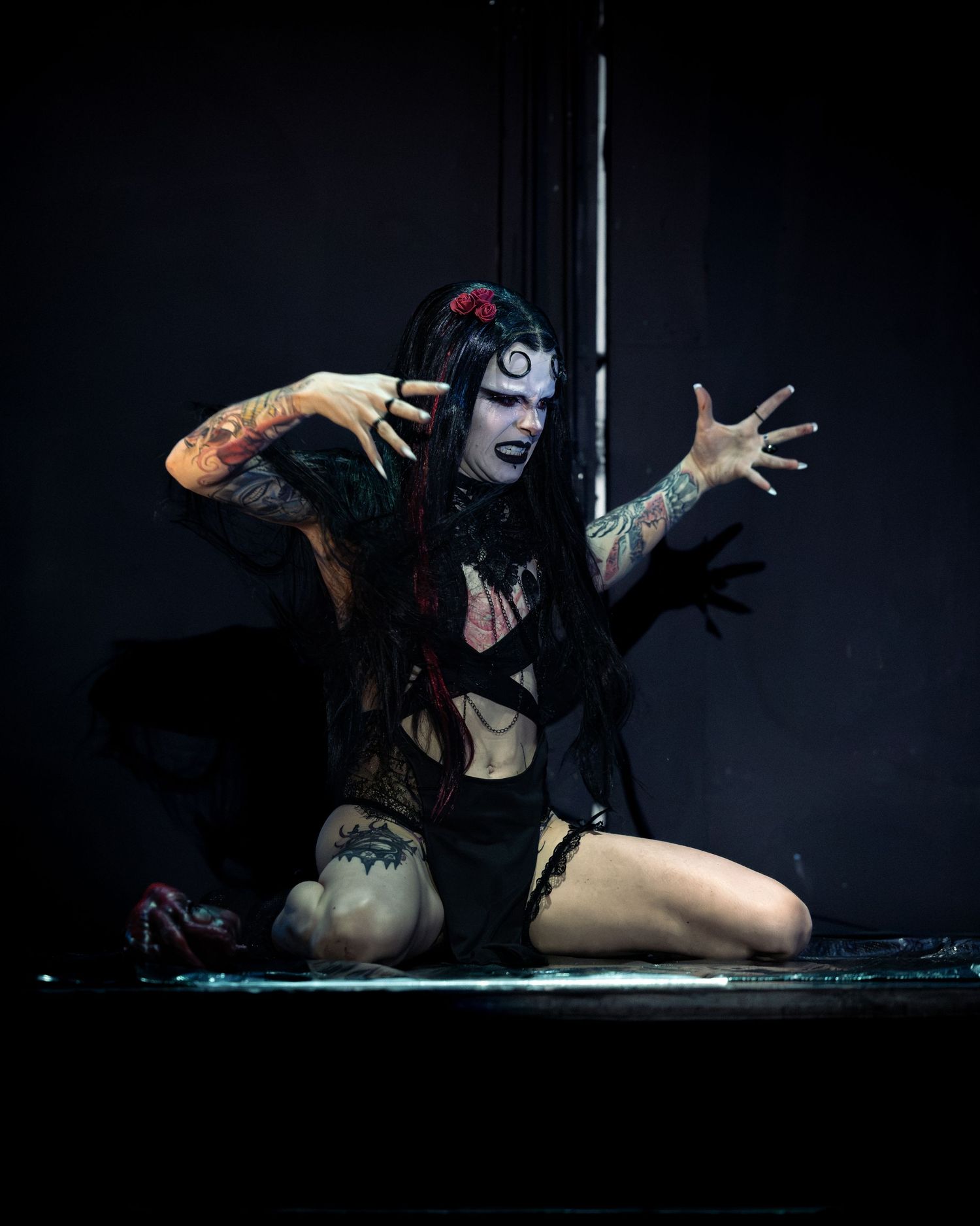Gothic horror performer with white face paint and dramatic makeup poses on stage floor with arms raised.