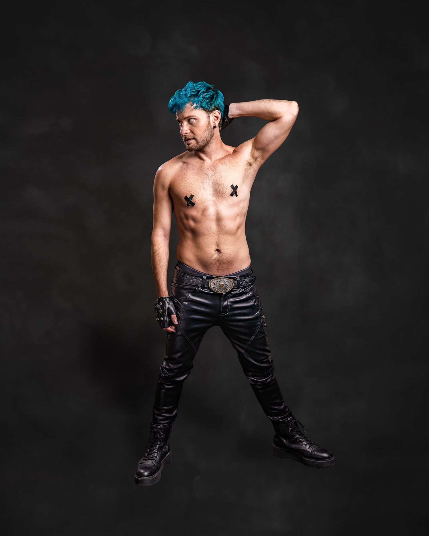 Shirtless man with teal hair wearing black leather pants and gloves poses confidently against dark studio background.