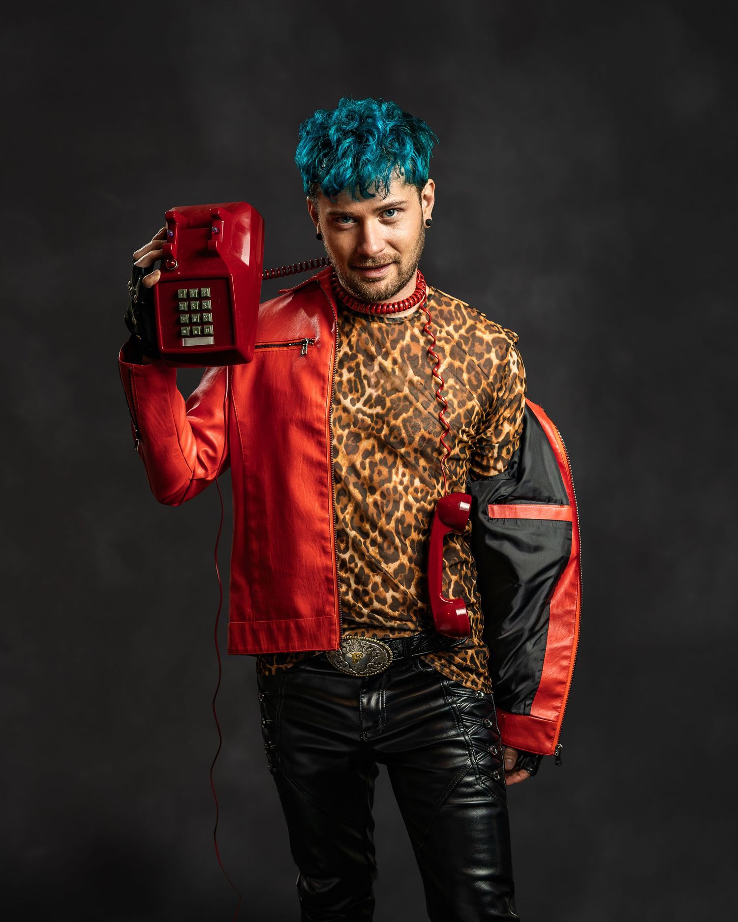 Man with blue hair balances red vintage telephone on shoulder, styled in red leather jacket and leopard print shirt.