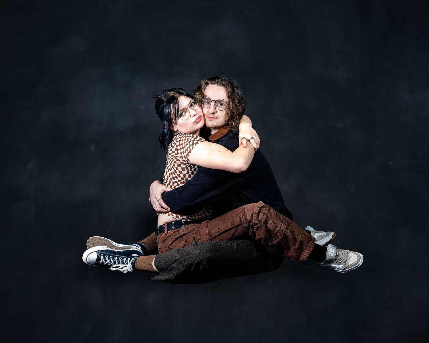 Two young people sitting on floor hugging in studio portrait against dark background.
