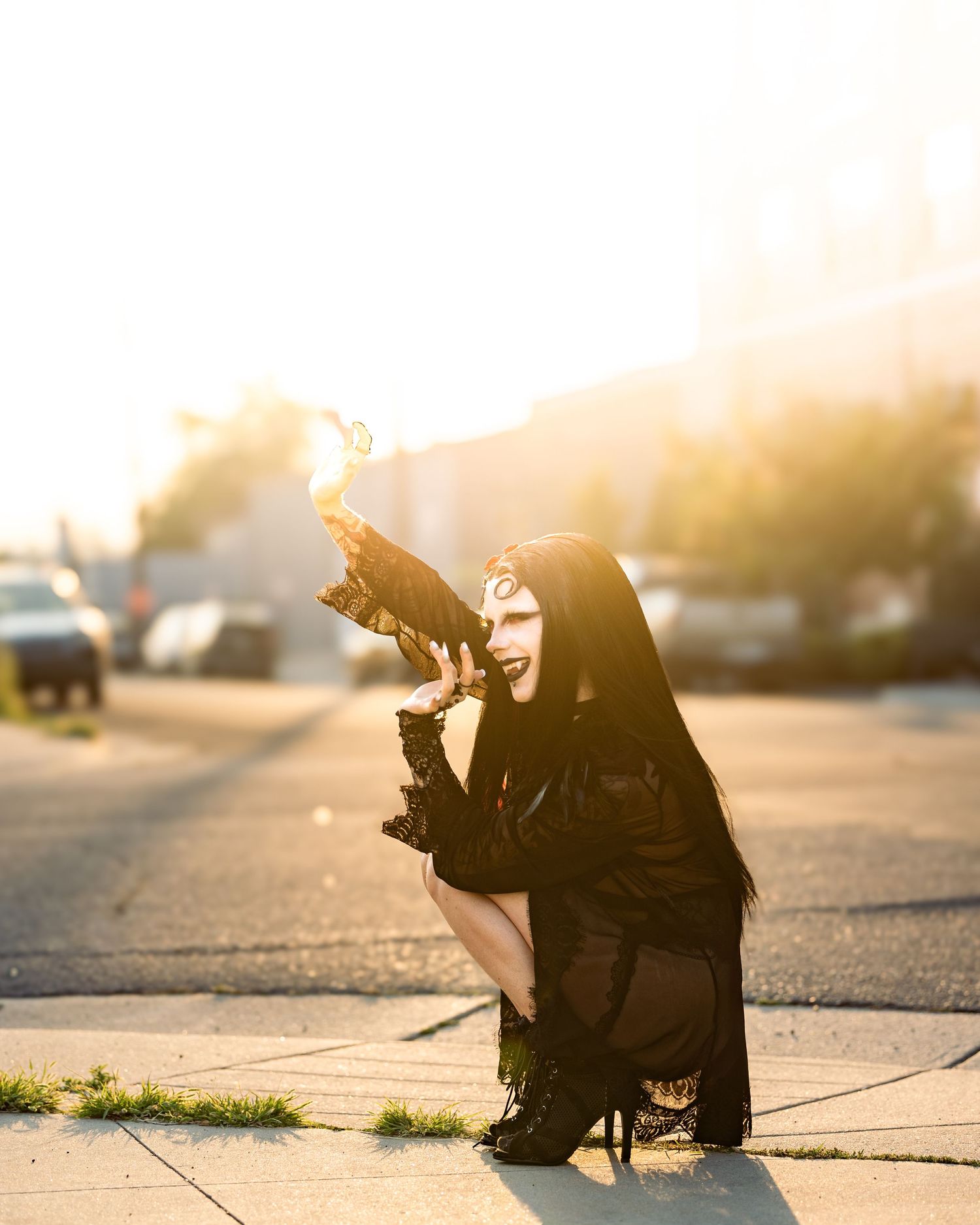 Gothic performer in black outfit kneels on sunlit street, dramatically raising a prop toward golden light.