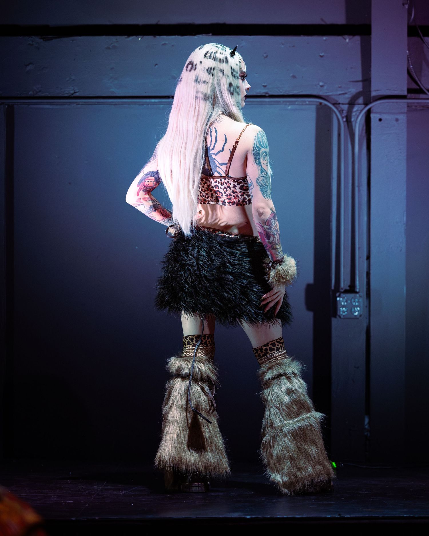 Heavily tattooed performer in leopard print crop top, black fur skirt, and fluffy leg warmers stands on stage.