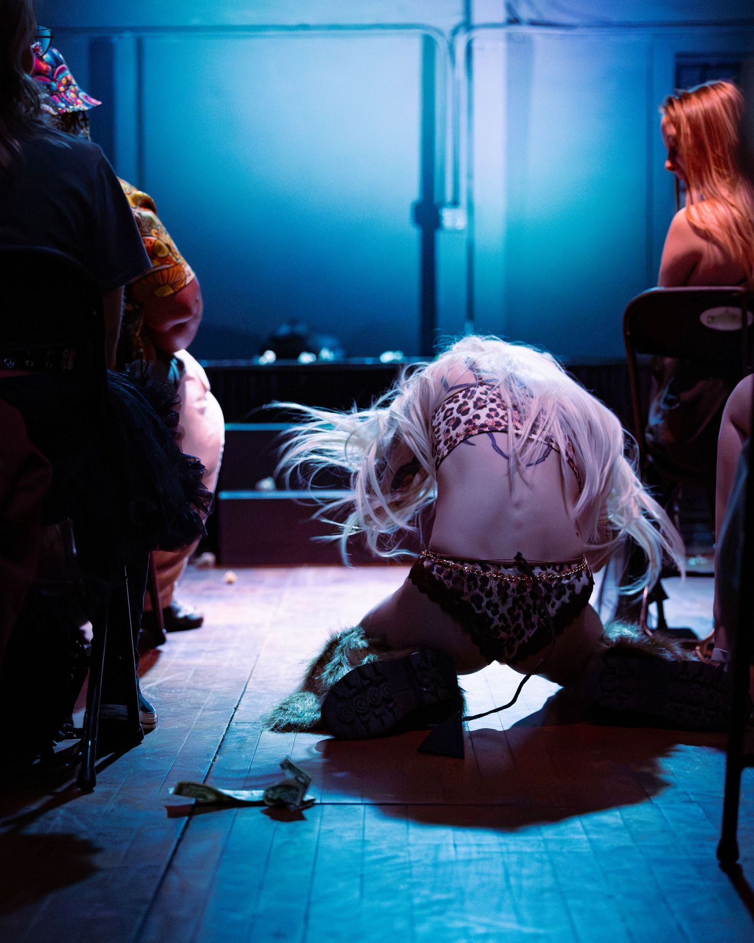Tattooed blonde performer in leopard print crouching on stage floor near seated audience.