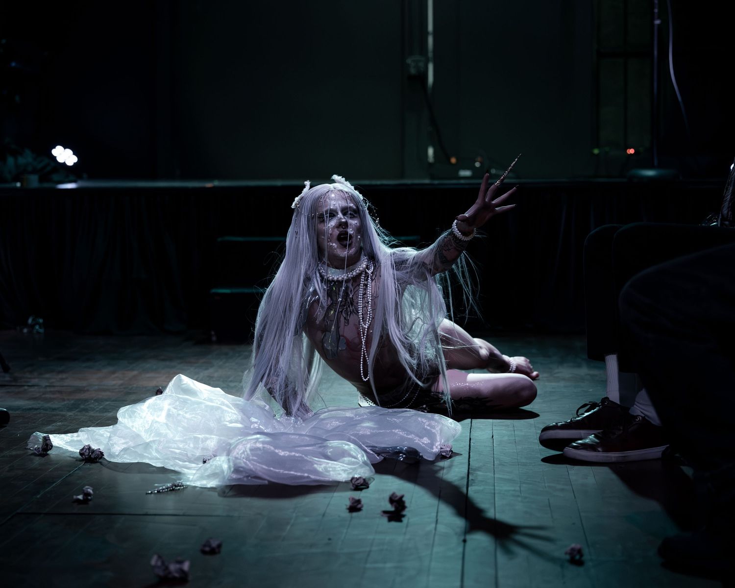 Eerie ghost bride costume with white dress and long hair, sitting on dark stage floor with dramatic pose.