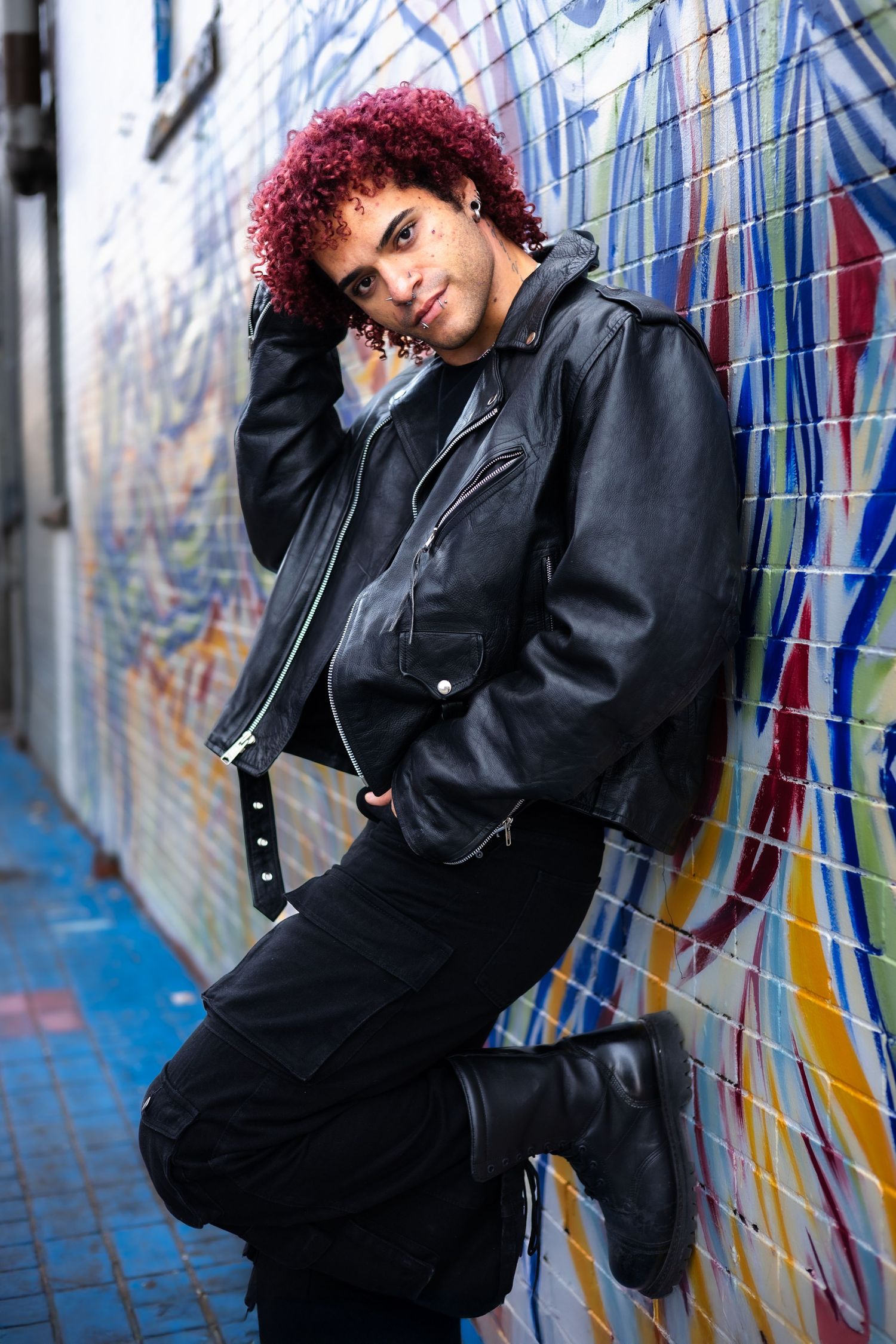 Person with red curls poses stylishly against blue and yellow street art.