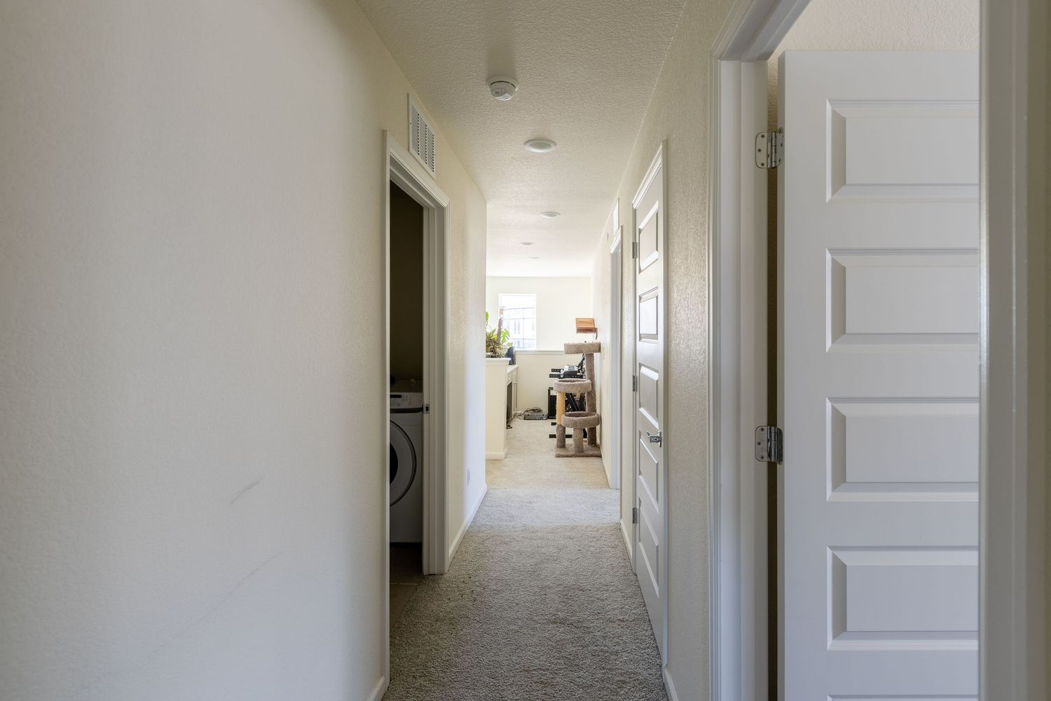 Bright narrow hallway with carpet flooring leading to laundry area, featuring white walls and interior doors.