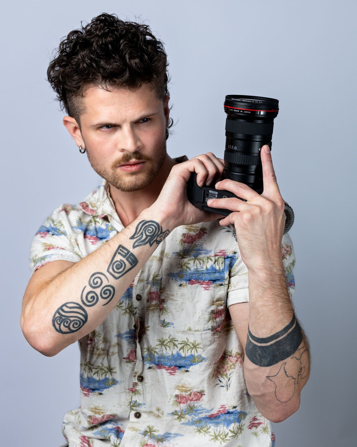 Tattooed male photographer holding a Canon camera, wearing a floral shirt, with curly hair and an ear piercing.