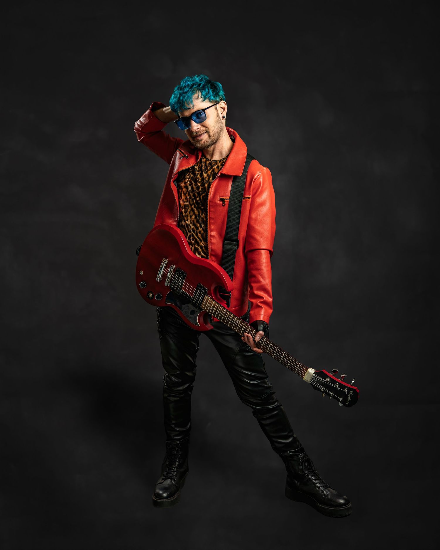 Rock musician with teal hair in a red leather jacket poses with a red electric guitar on dark background.