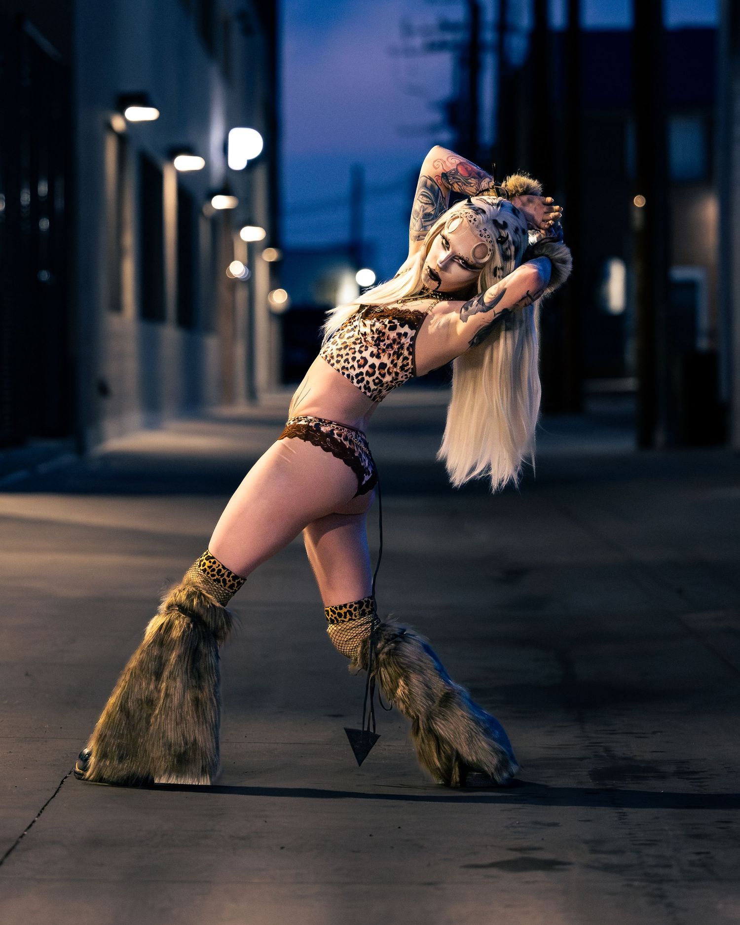 Woman in leopard print outfit and fur boots poses dramatically on an urban street at dusk.