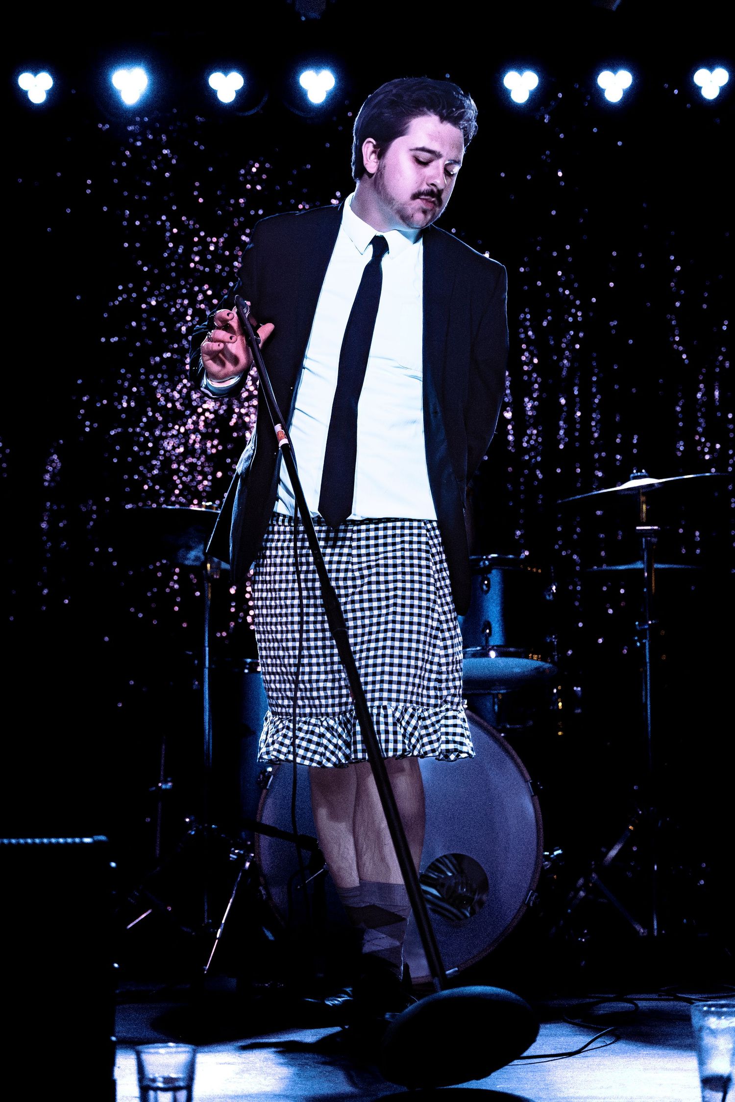 Male performer in black blazer, tie, and checkered skirt stands at microphone under sparkly stage lights.