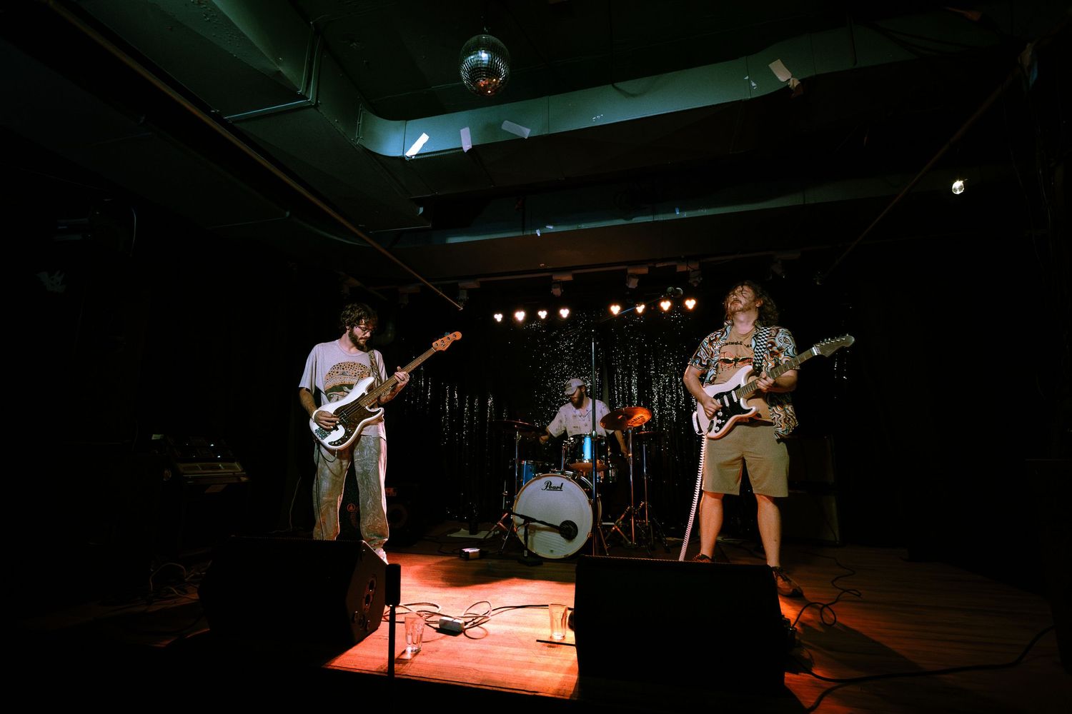 Three-piece rock band performs on a small indoor stage with dramatic lighting and disco ball.
