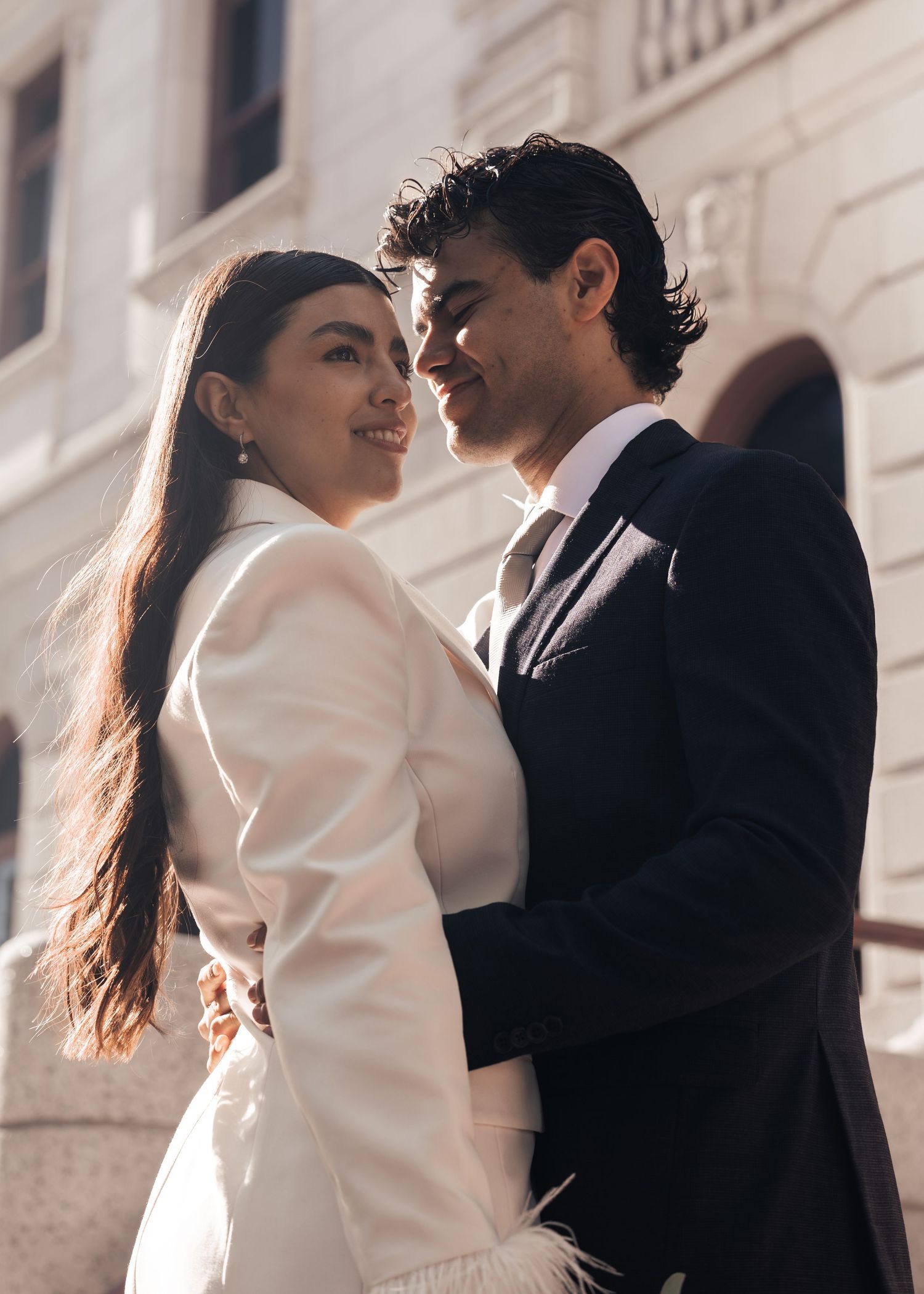 Elegant couple in wedding attire embracing outdoors, woman in white blazer and man in dark suit, smiling romantically.