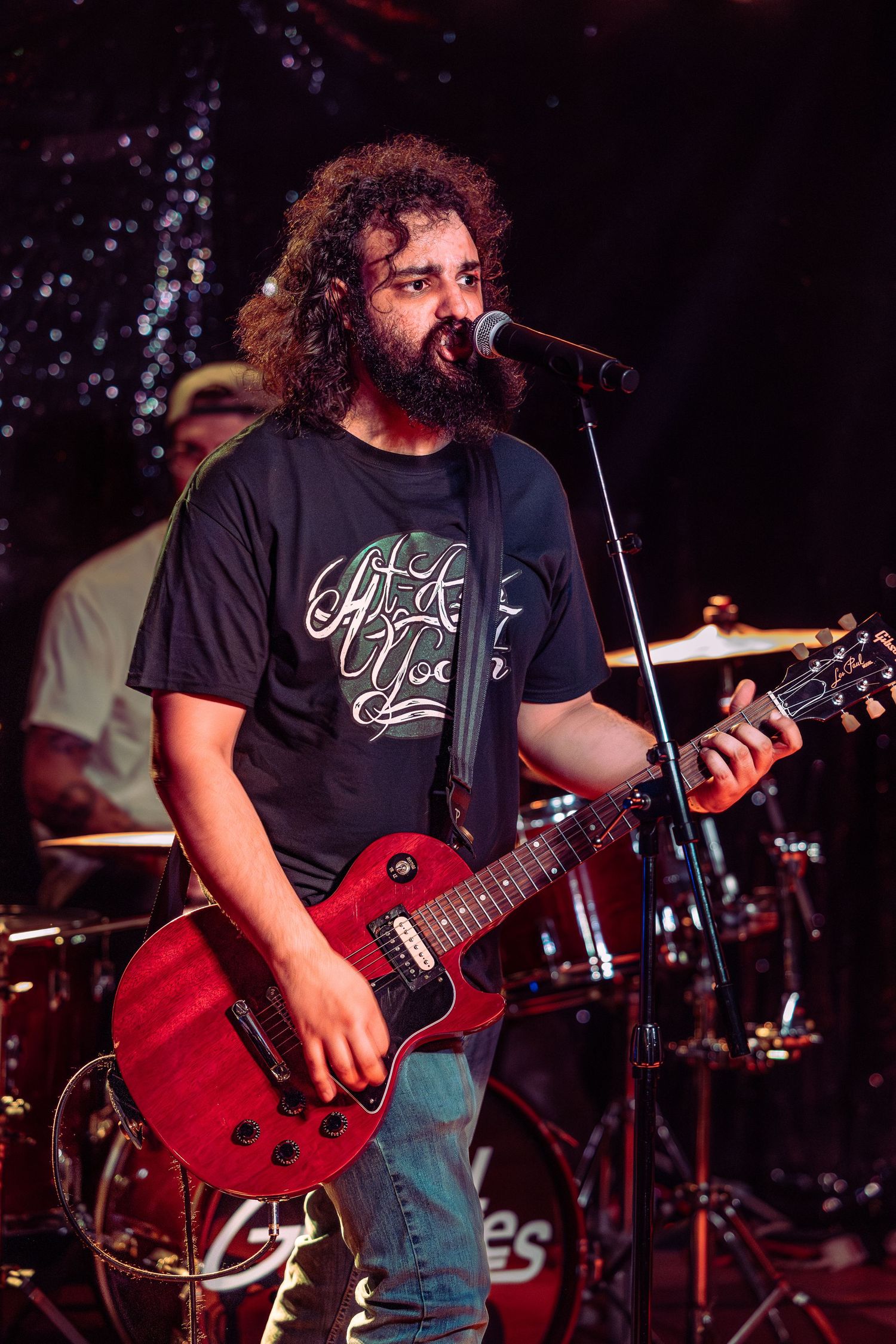 Curly-haired bearded musician singing and playing red Gibson Les Paul guitar under warm stage lighting.