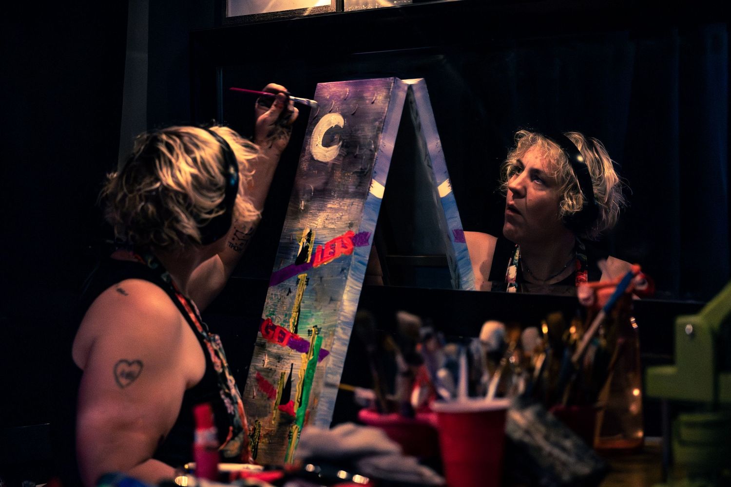 Artist painting colorful canvas on easel in dark studio with brushes and supplies visible nearby.