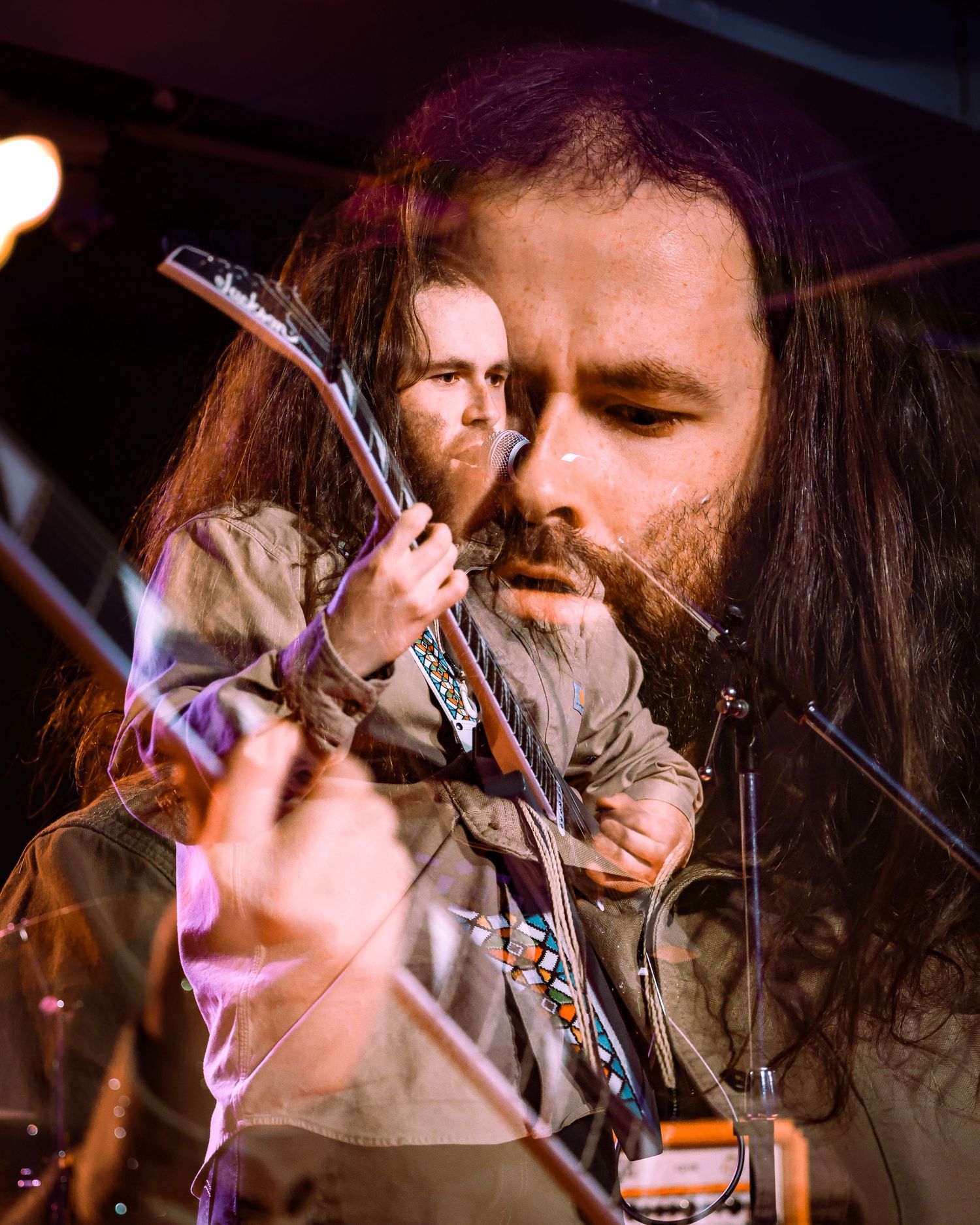 Long-haired bearded guitarist playing electric guitar under warm stage lights at live show.