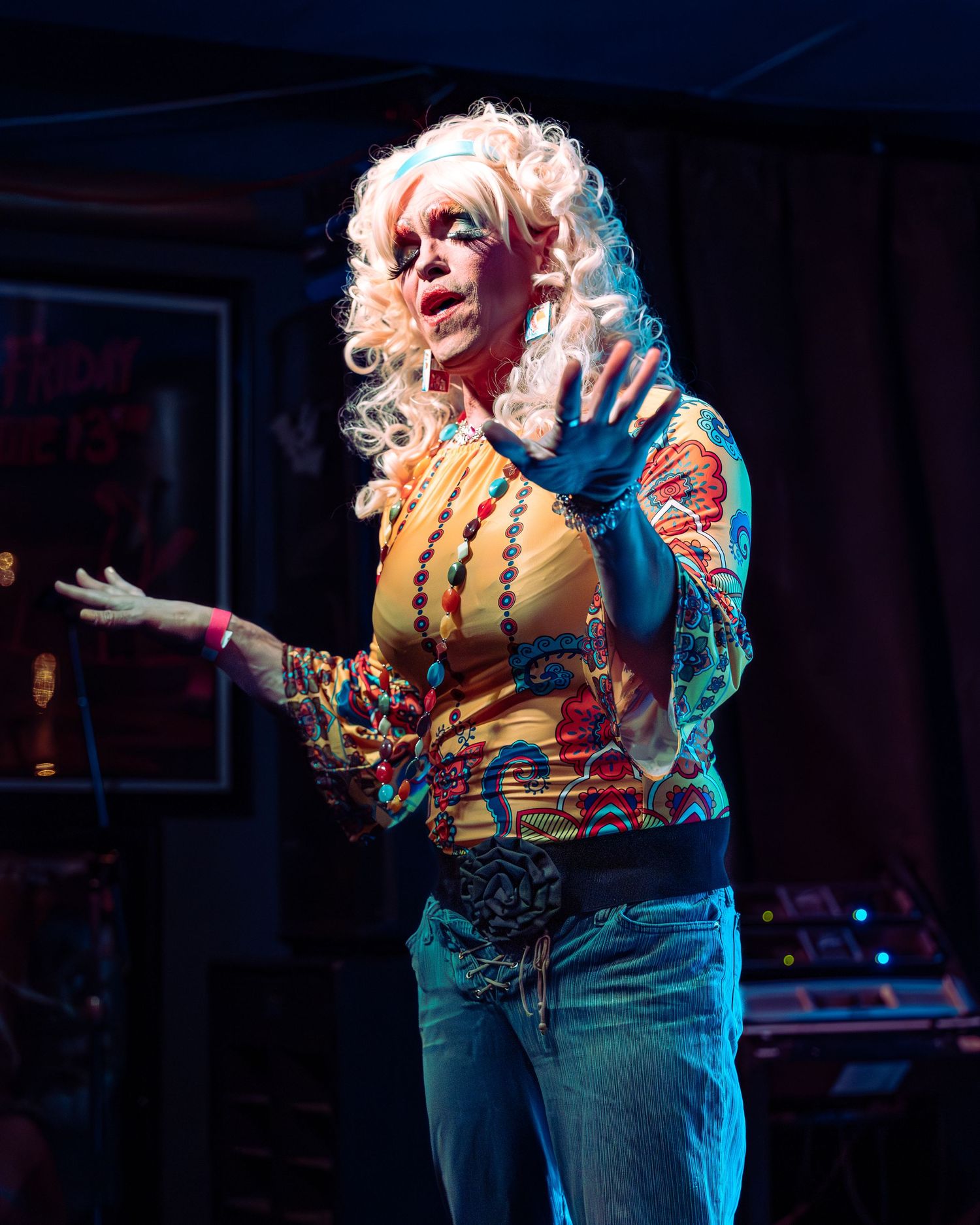 Drag queen in white curly wig and vibrant floral outfit gestures expressively on stage.