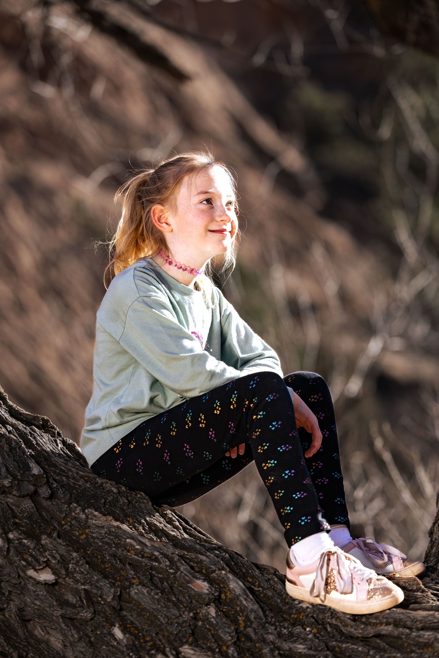 A smiling young girl with blonde hair sits on a tree trunk outdoors, wearing a mint sweatshirt and patterned leggings.