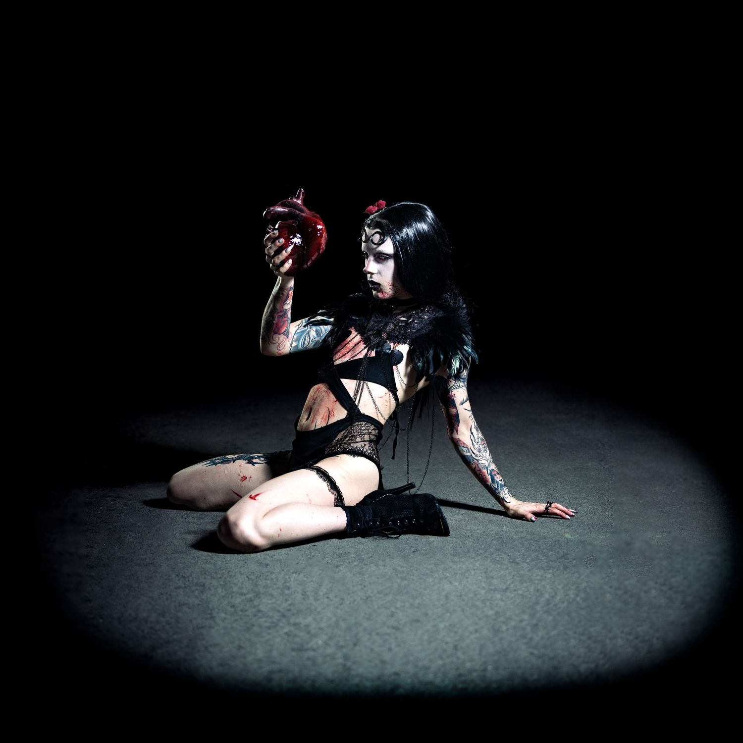 Tattooed goth woman in black lingerie kneeling dramatically in spotlight holding red heart prop.