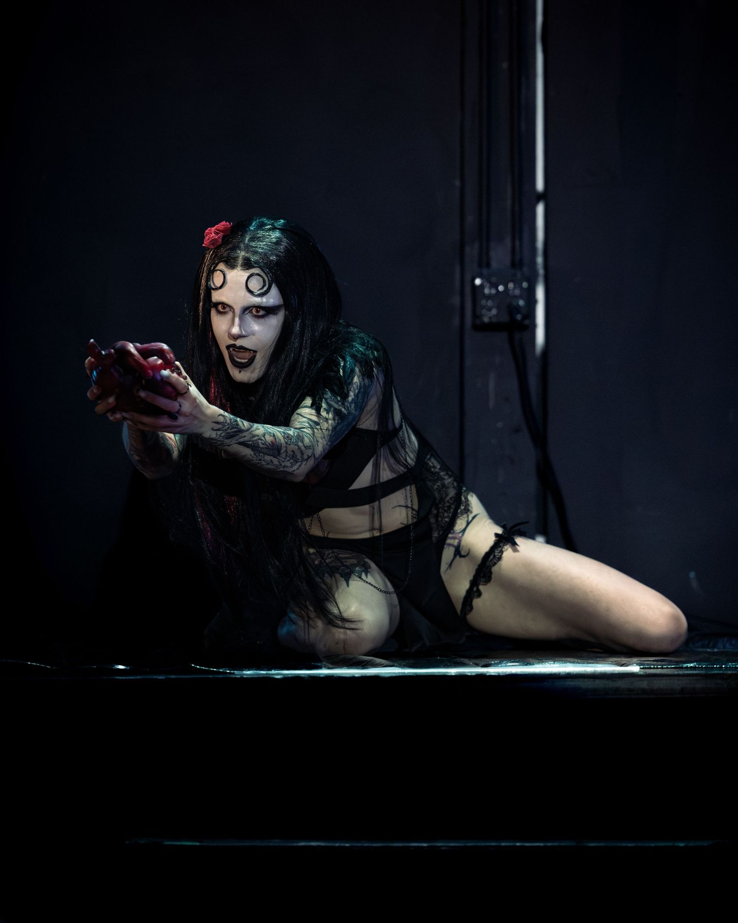 Dramatic gothic performer with face paint and tattoos crouching on wet stage holding red heart prop.