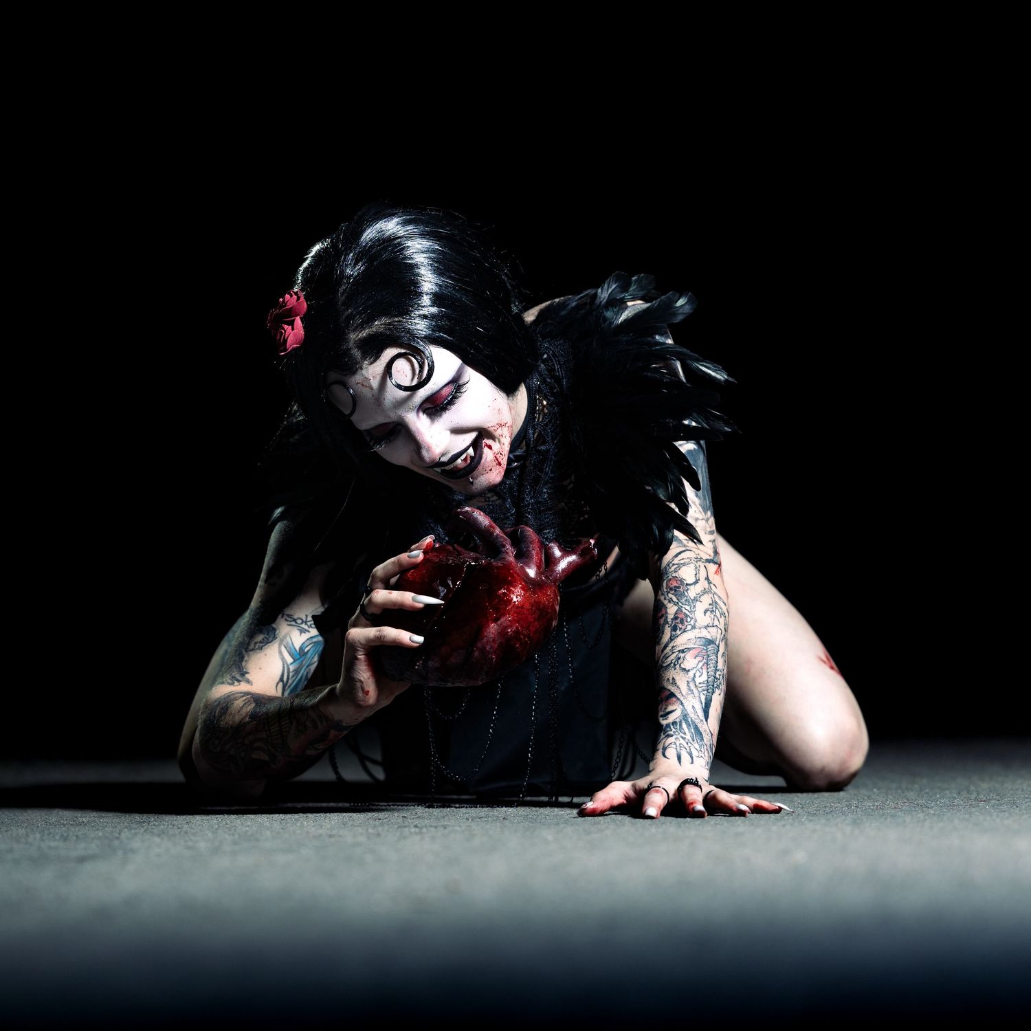 Gothic woman with black hair, dramatic white face paint, tattoos, and dark feathers crawls on ground in dark studio setting.