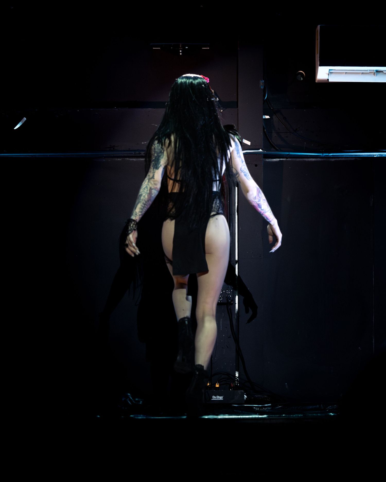 Tattooed gothic performer in black lingerie and cape standing on dark stage from behind.
