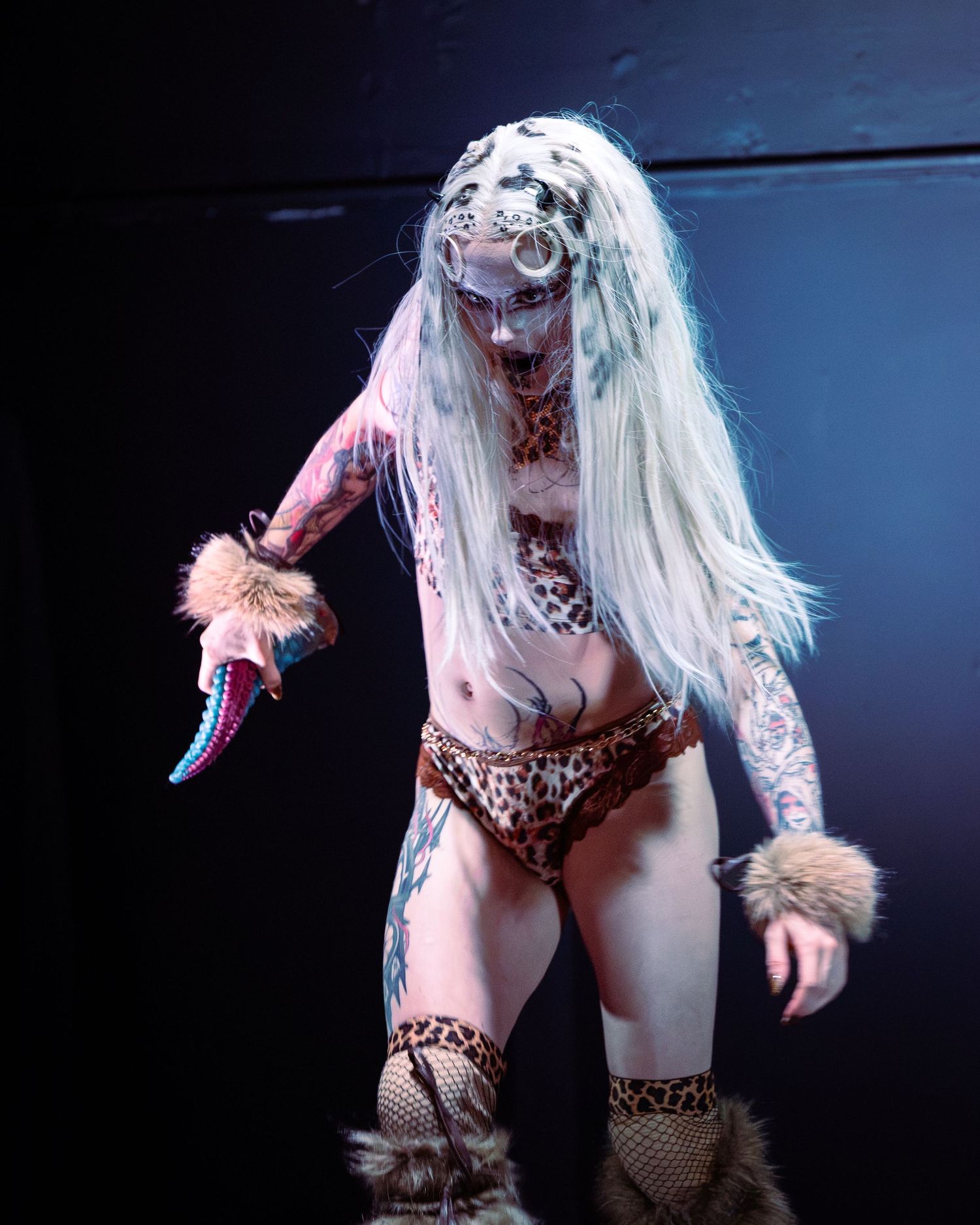 Tattooed burlesque performer in leopard print bikini holding colorful prop with dalmatian face paint.