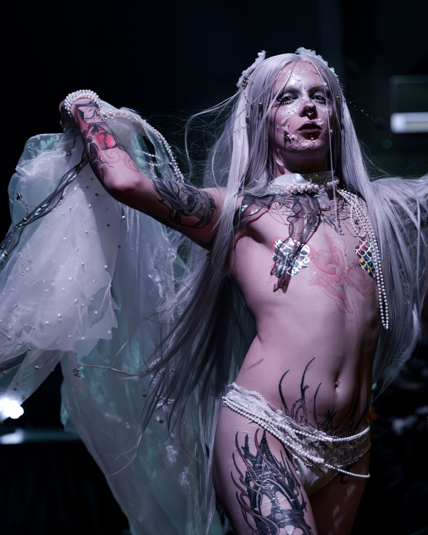 Dramatic performer with silver hair, body paint, tattoos, and sheer white costume on dark stage.