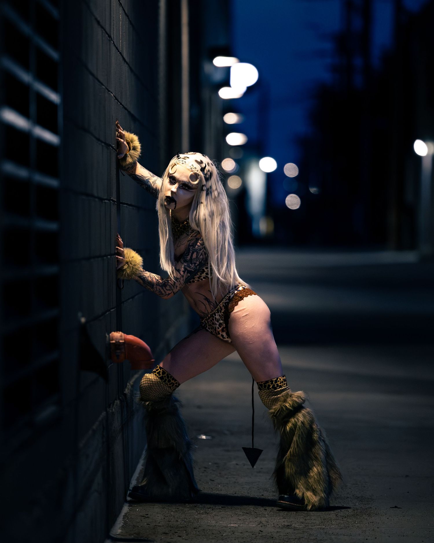 Costumed tattooed performer in cheetah mask and fur boots leans against brick wall at night.