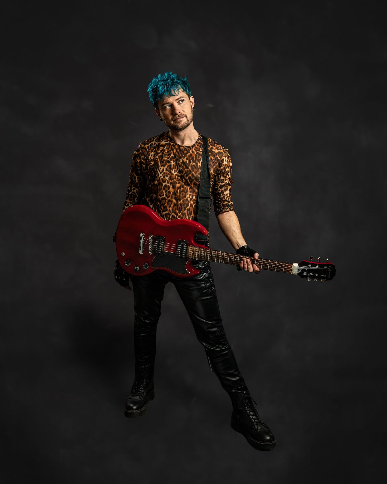 Musician with teal hair in leopard print shirt holding red electric guitar against dark background.
