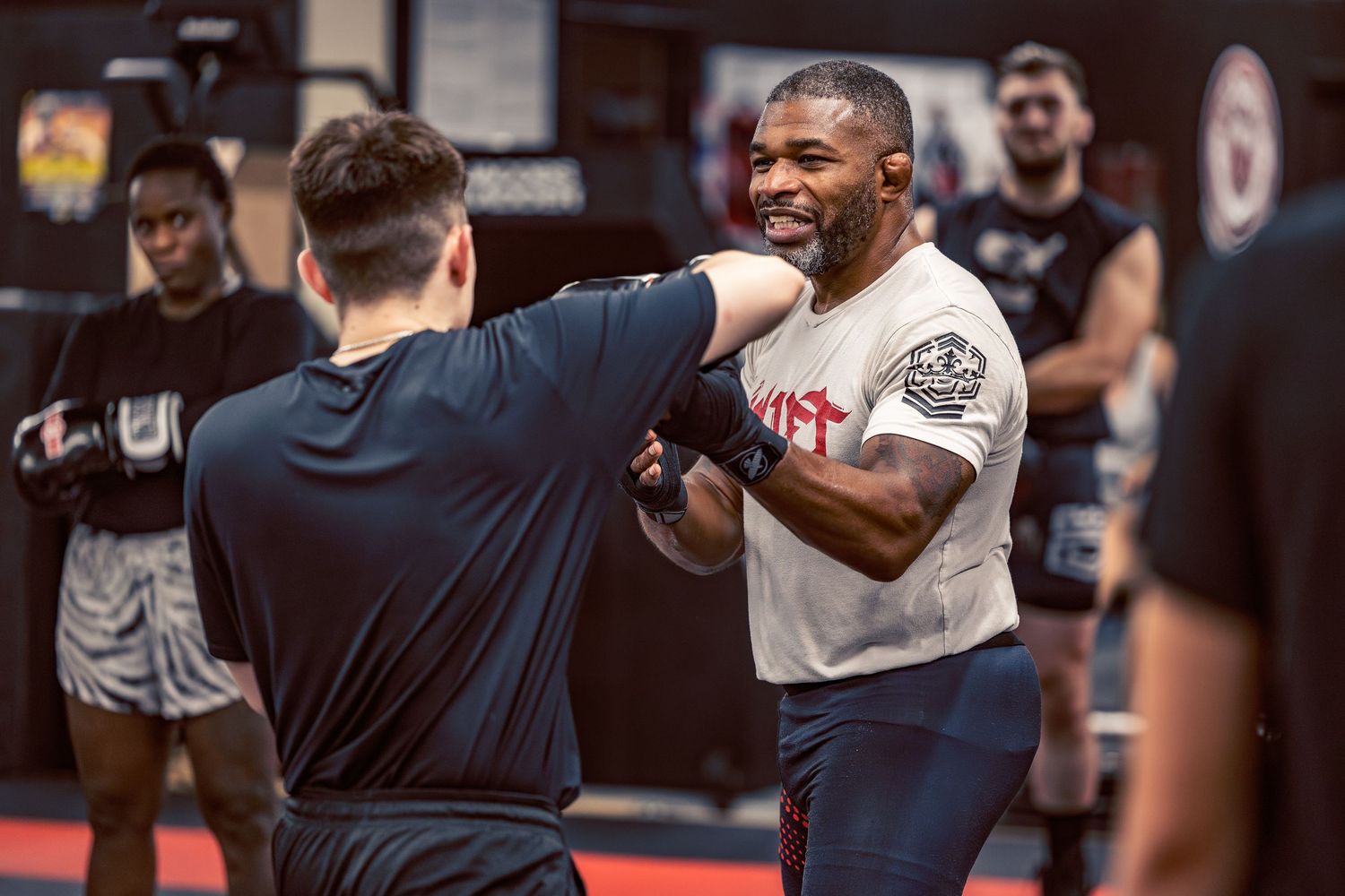 Mixed martial arts fighters practicing sparring techniques in a gym with red mats and championship belts on display.