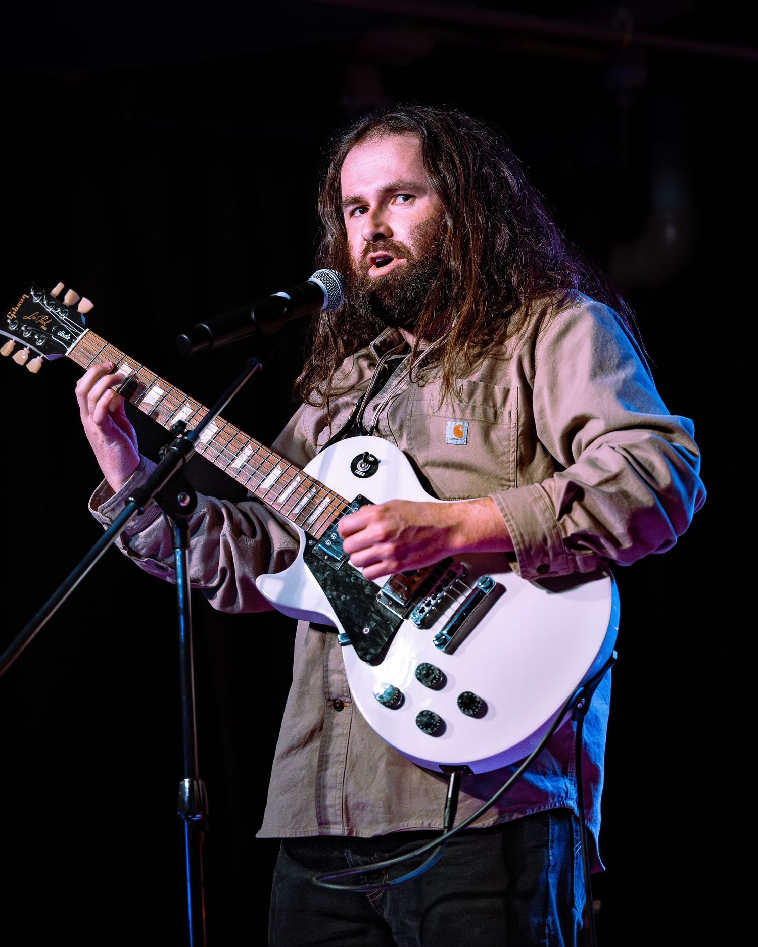 Long-haired bearded musician playing a white electric guitar at a microphone on stage.