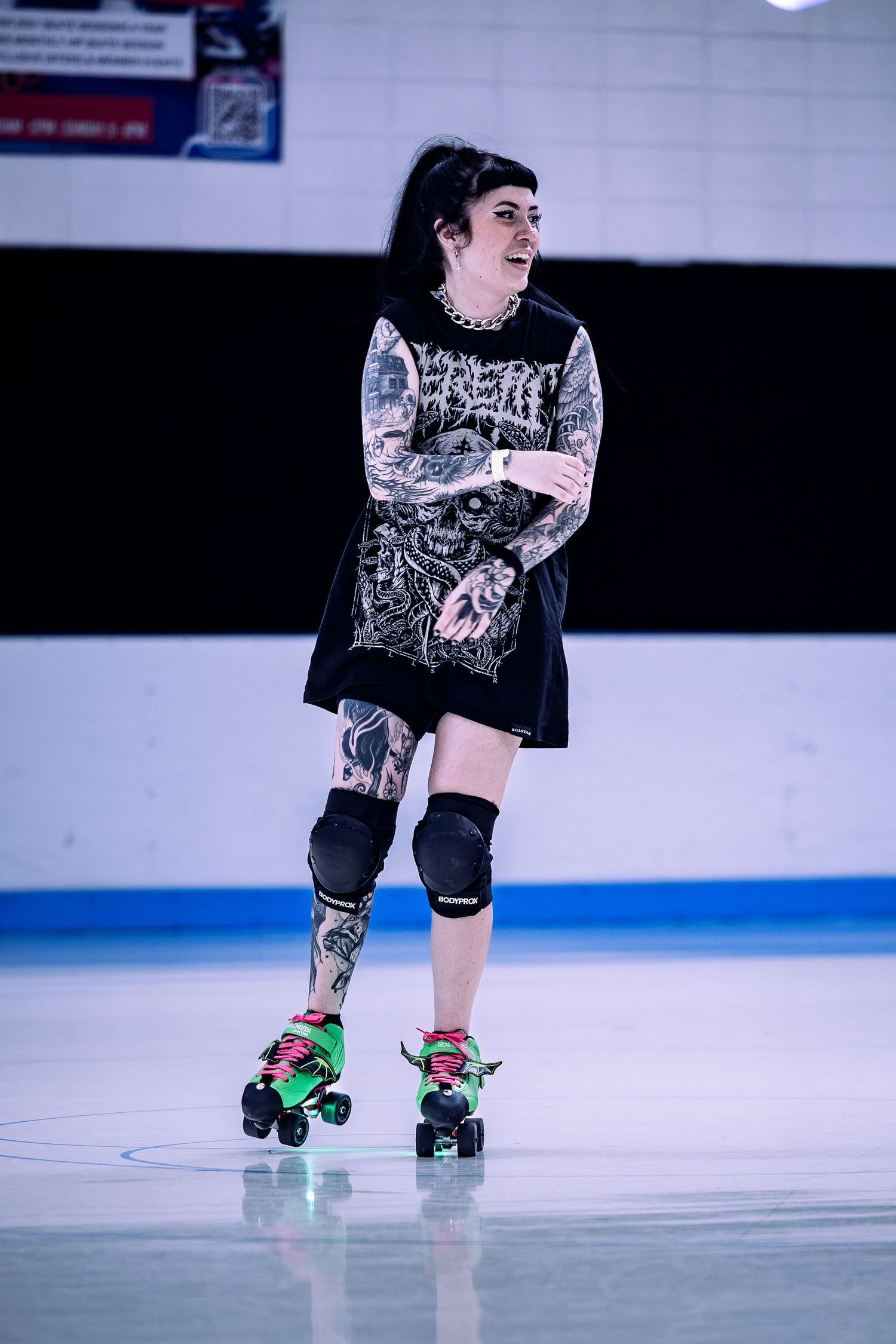 Tattooed woman in black outfit and knee pads skating on roller skates at an indoor roller rink.