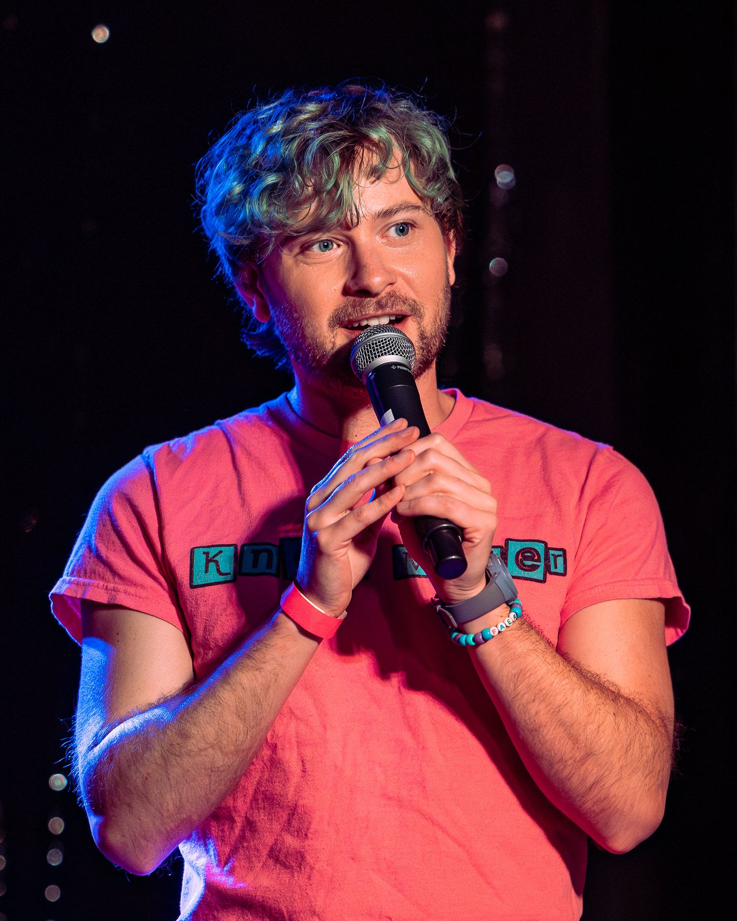 Young man in pink t-shirt holding microphone performing on stage under blue and pink lights.