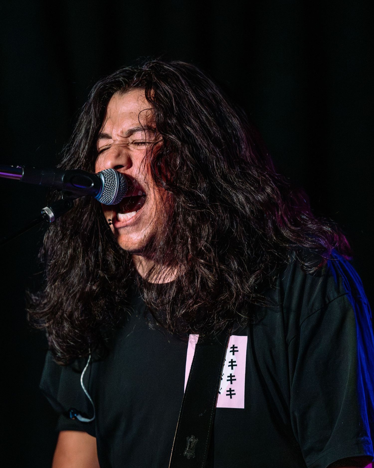 Long curly-haired male vocalist sings intensely into microphone wearing black Japanese text t-shirt on stage.
