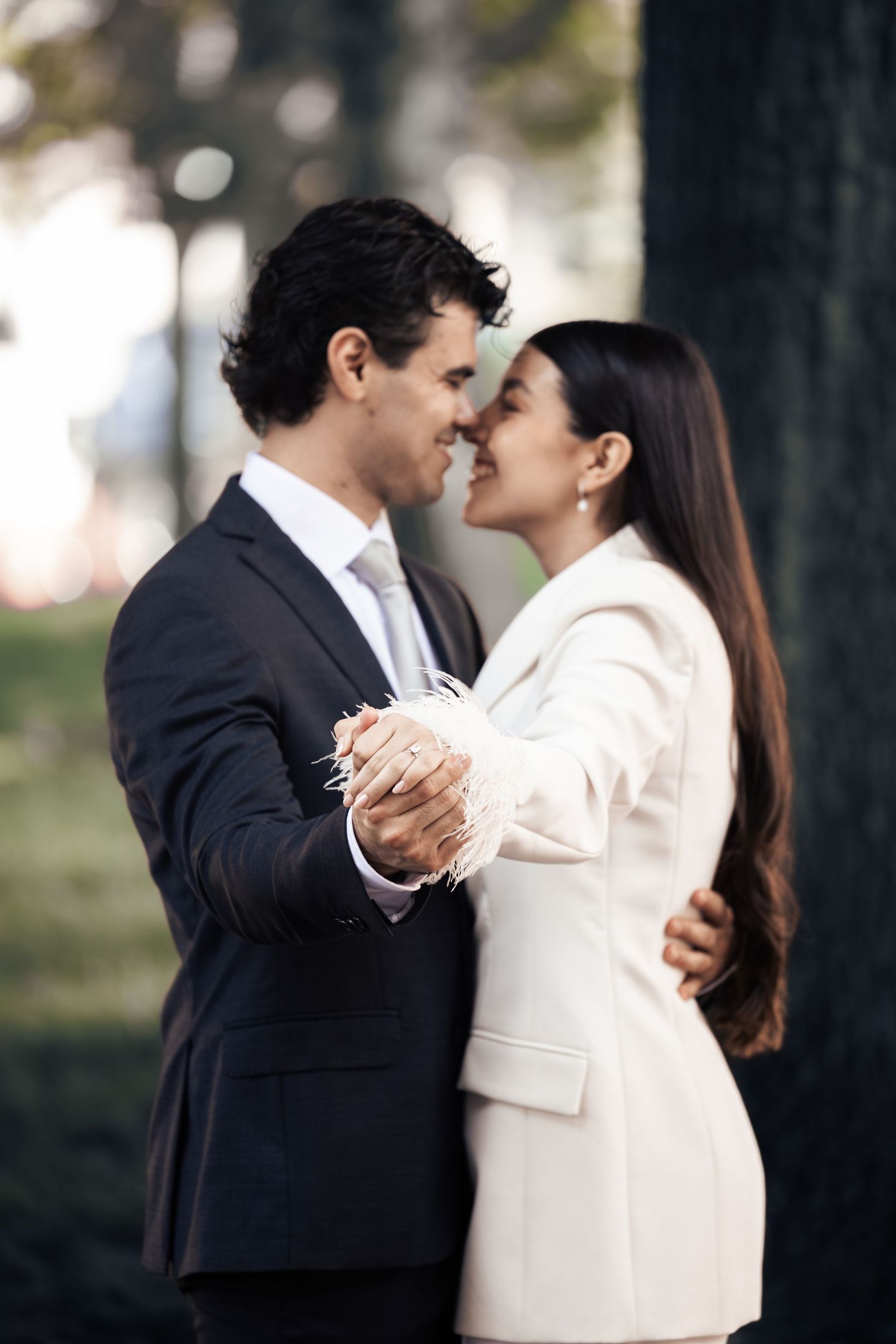 A couple in formal attire shares a romantic moment, nearly kissing outdoors near a tree.