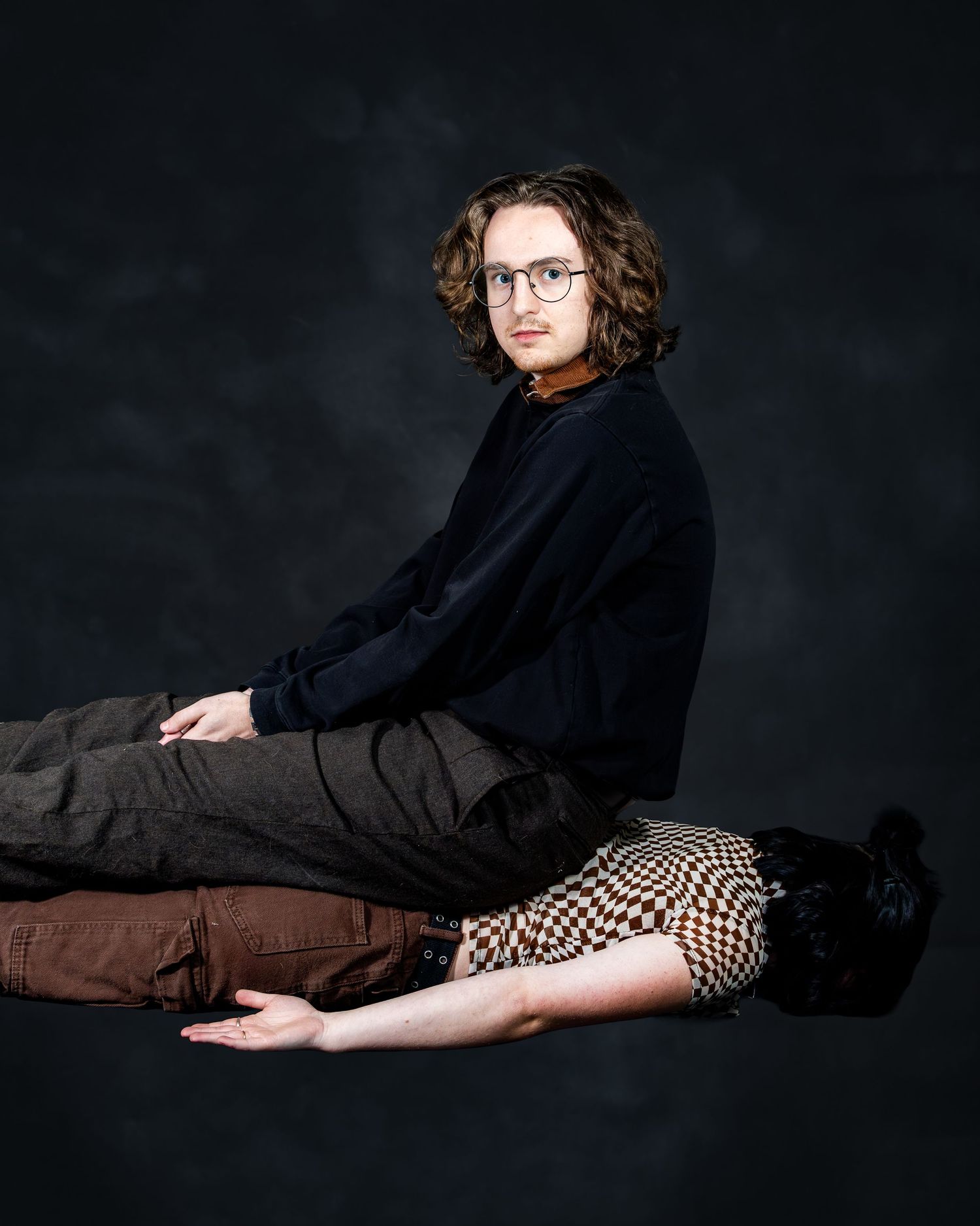 Young man with curly hair and glasses sits on a dark studio floor in a navy sweater and gray pants.