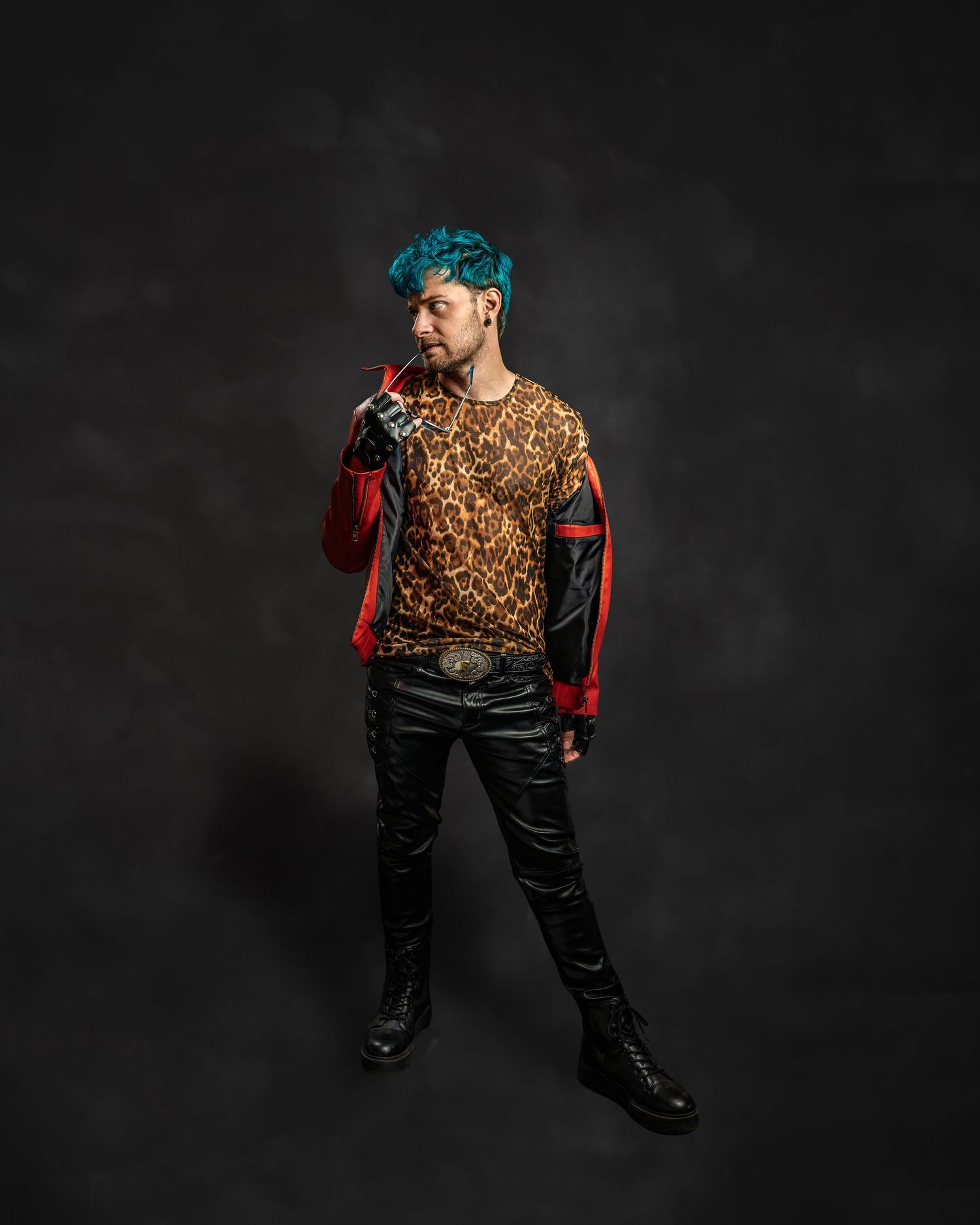 Person with teal hair wearing leopard print shirt, leather pants, and boots poses against dark background.