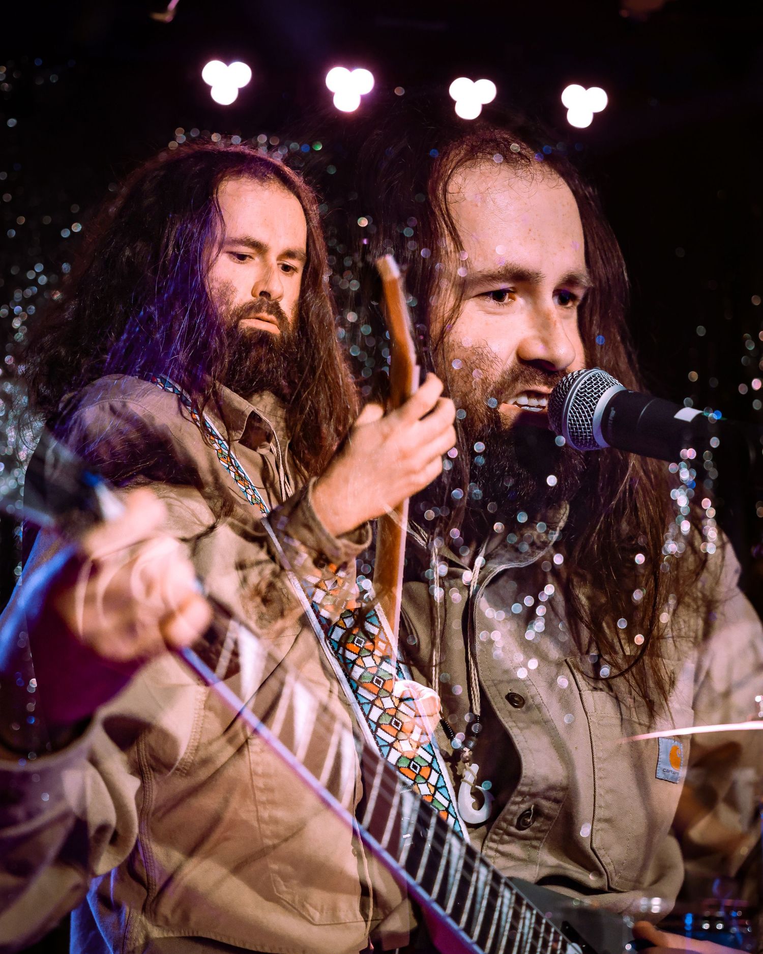 Two long-haired bearded musicians perform on stage, one singing into mic and one playing guitar.