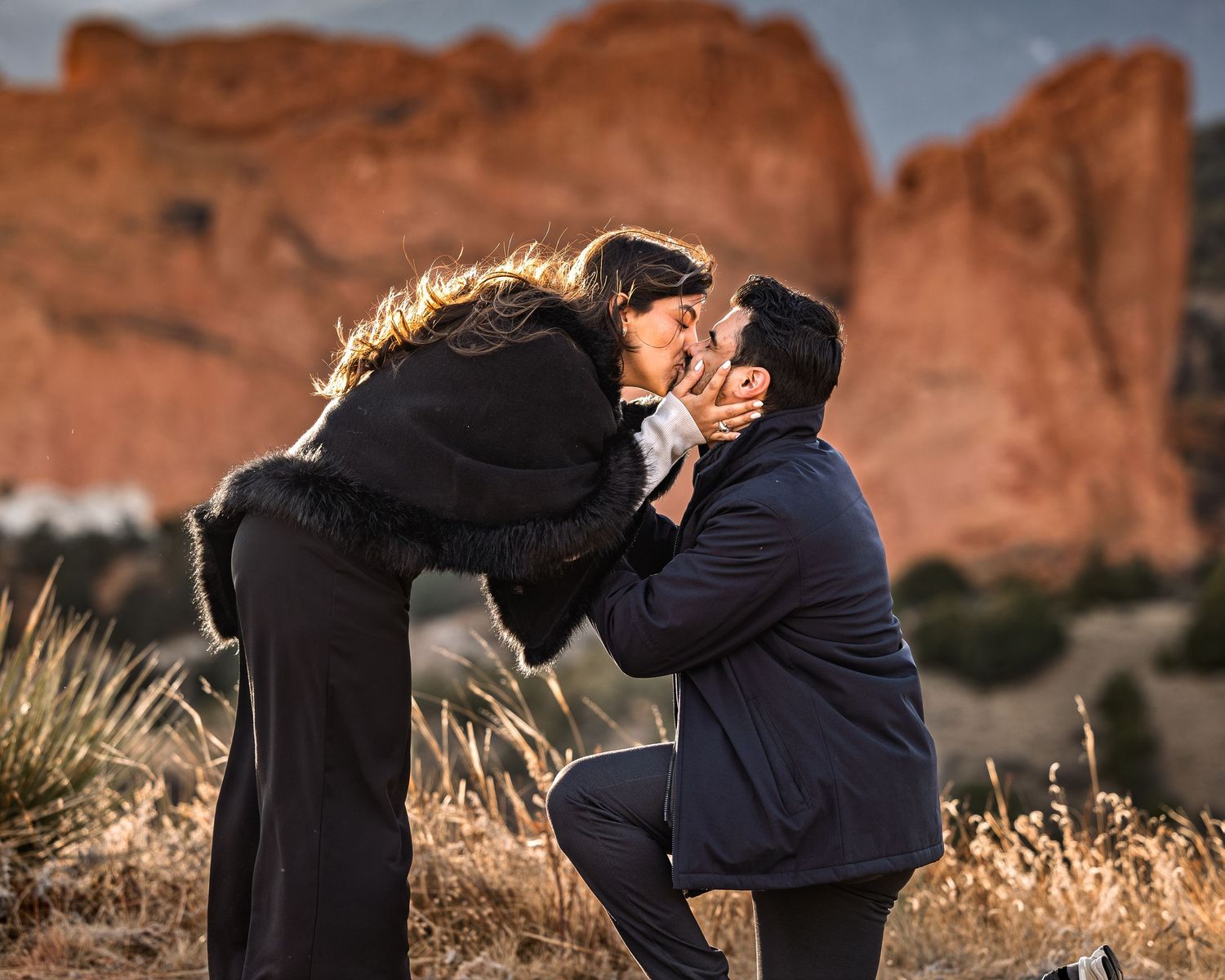 A couple embraces against a backdrop of red rock formations during golden hour at sunset.