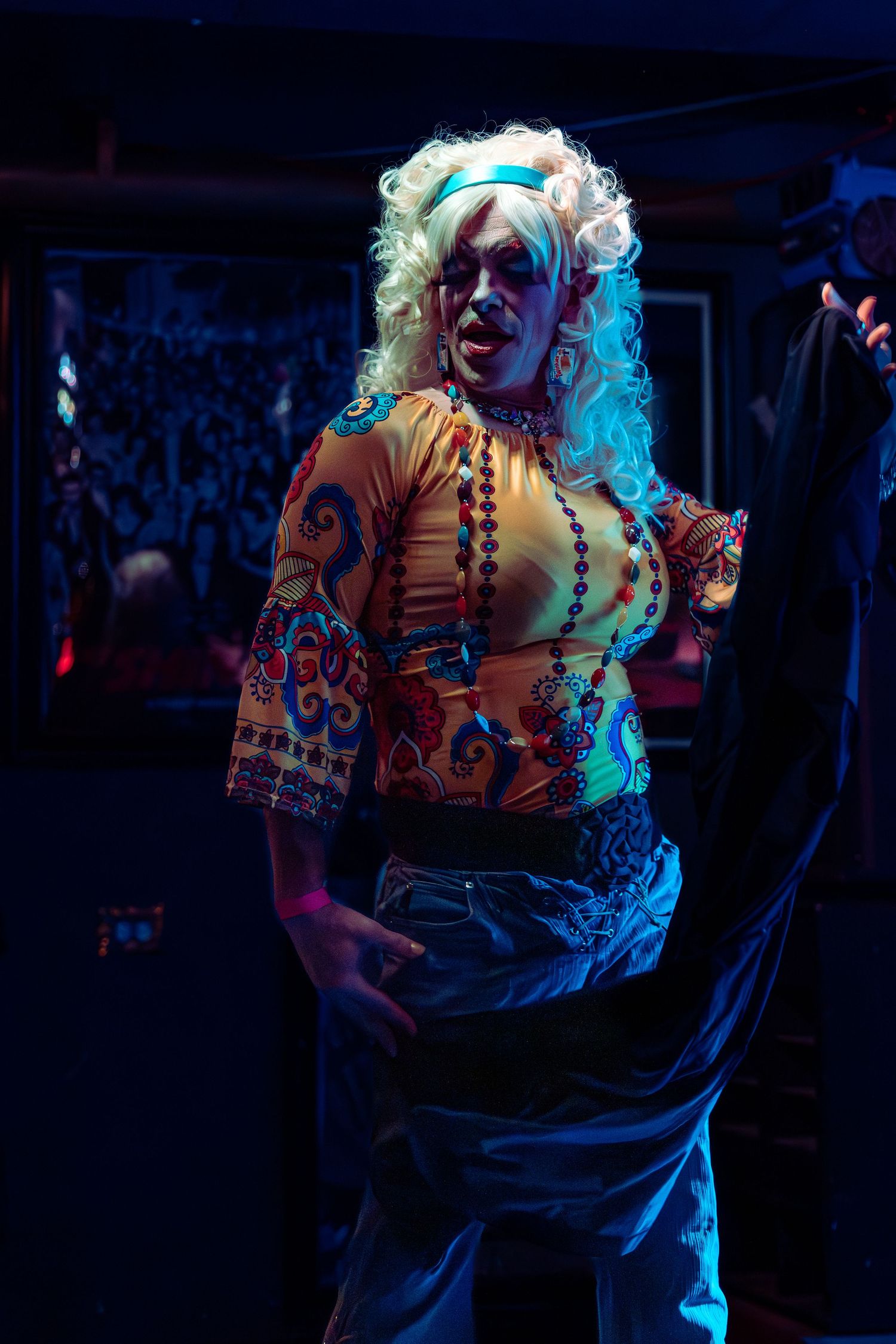 Drag performer in blonde wig and colorful patterned top poses on stage under blue and orange lights.