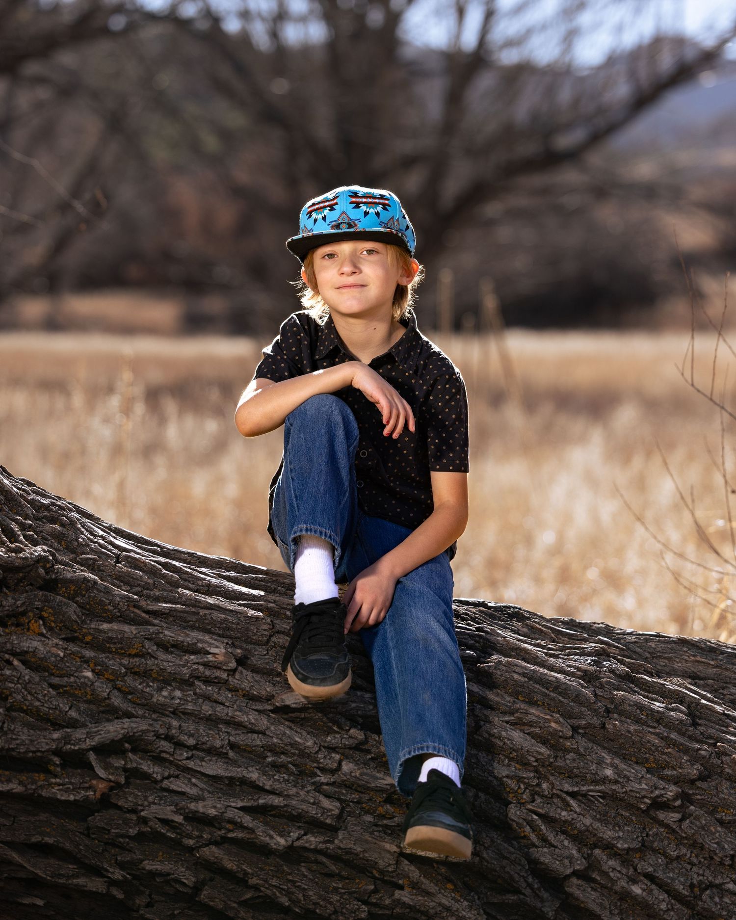 A young boy in jeans and a blue cap sits smiling on a fallen tree trunk in a golden field.