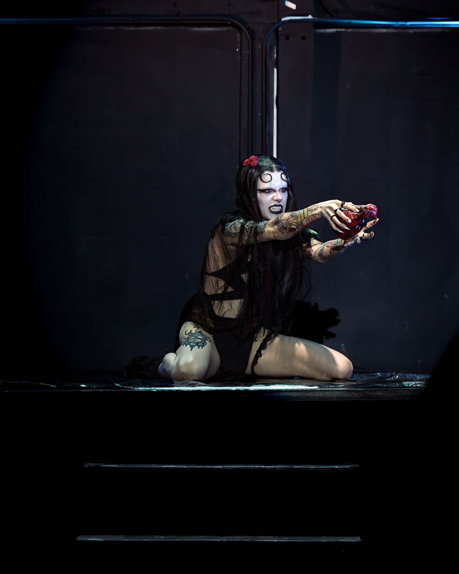 Gothic tattooed performer in sheer black dress kneeling on stage holding red heart in dark setting.